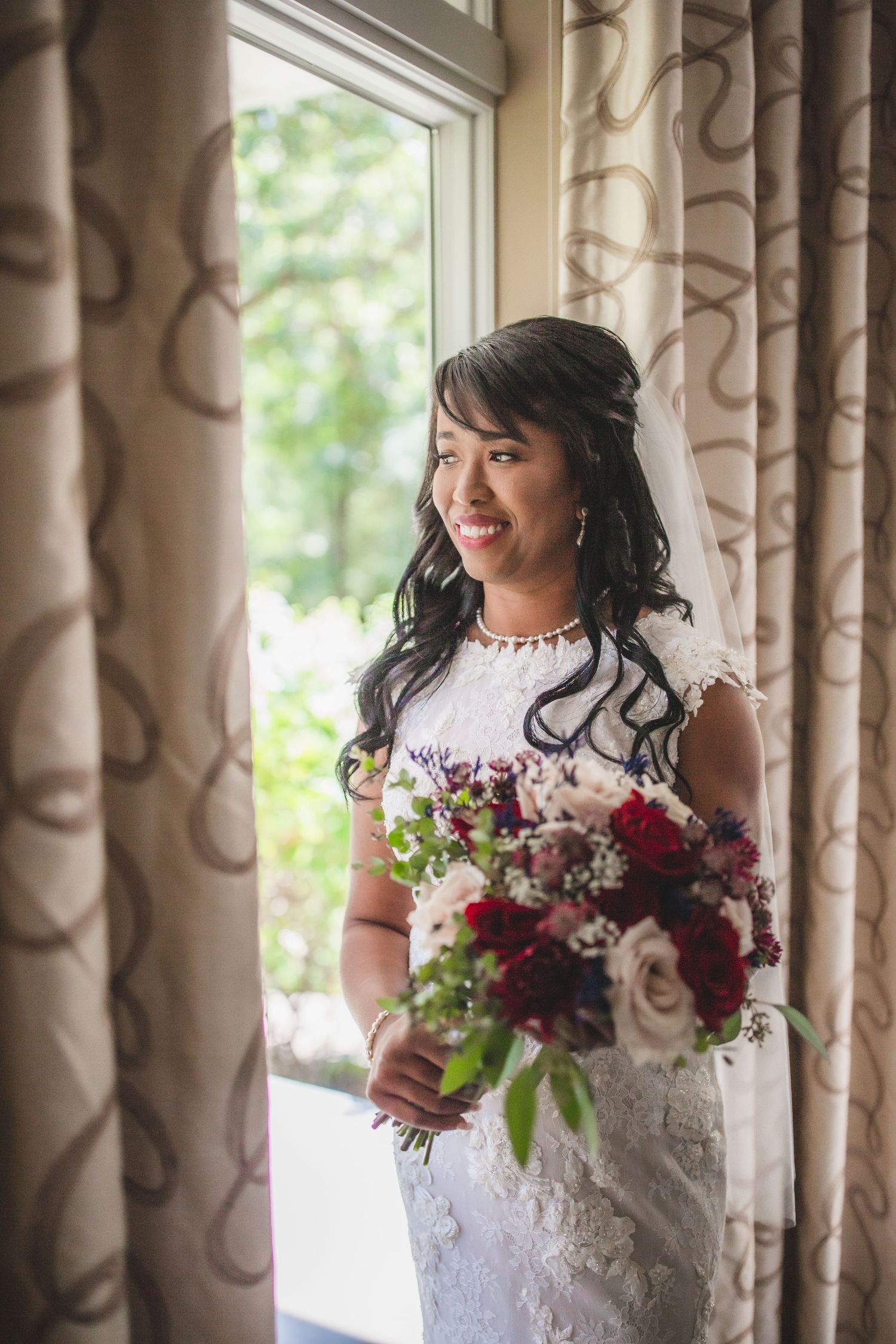 A bride smiles while holding her wedding bouquet next to a bright window with elegant curtains.