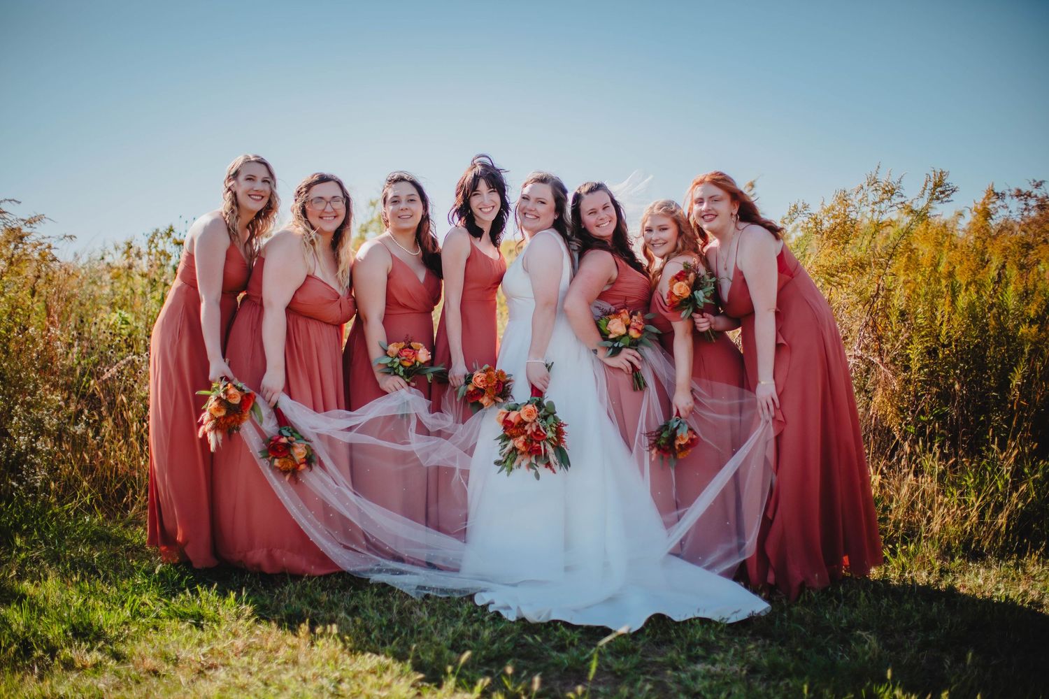 Bridal party in flowing coral dresses poses with bride's veil creating movement in outdoor setting.