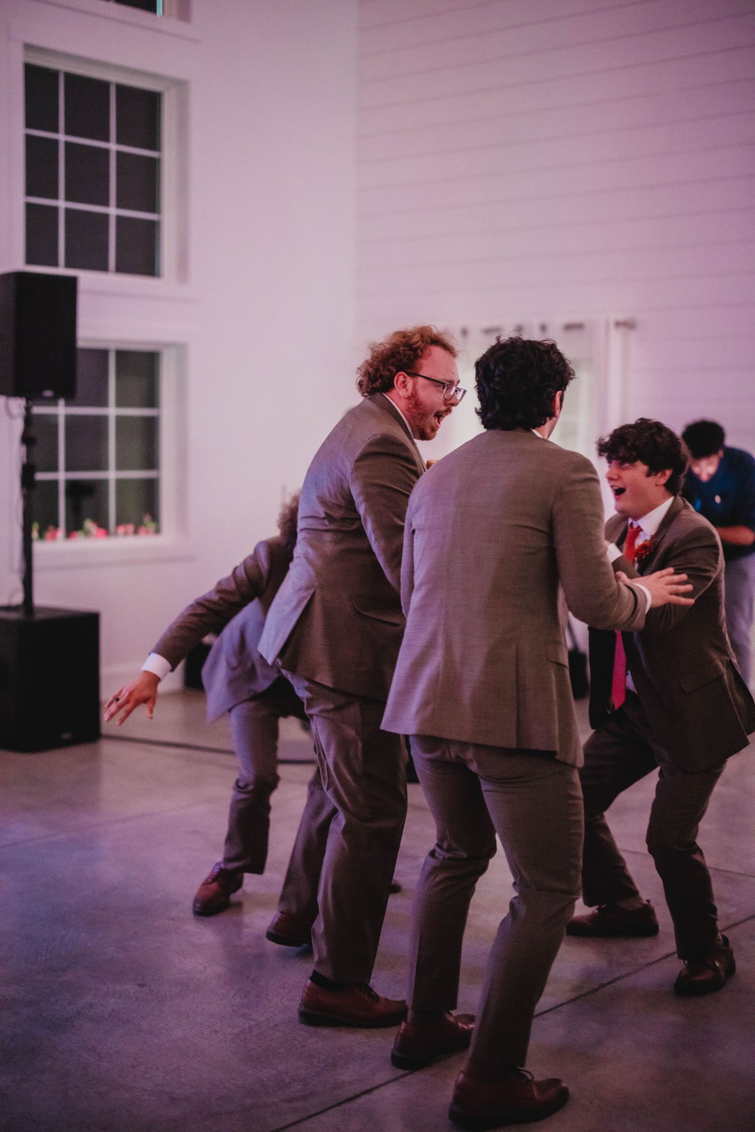 Guests dancing energetically in suits at an indoor reception with white walls and large windows.