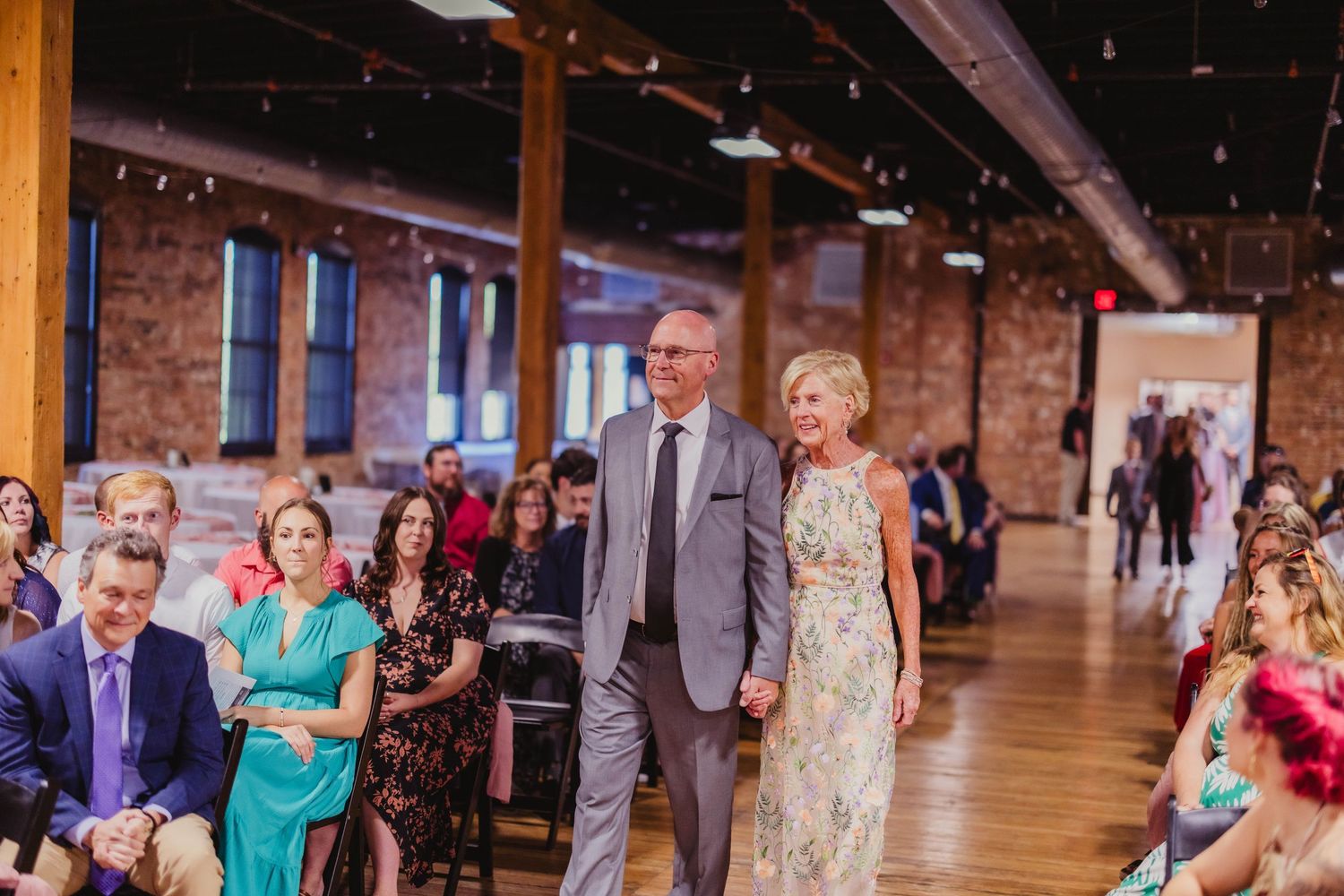 A couple walks down the aisle during a wedding ceremony in a rustic brick-walled venue.