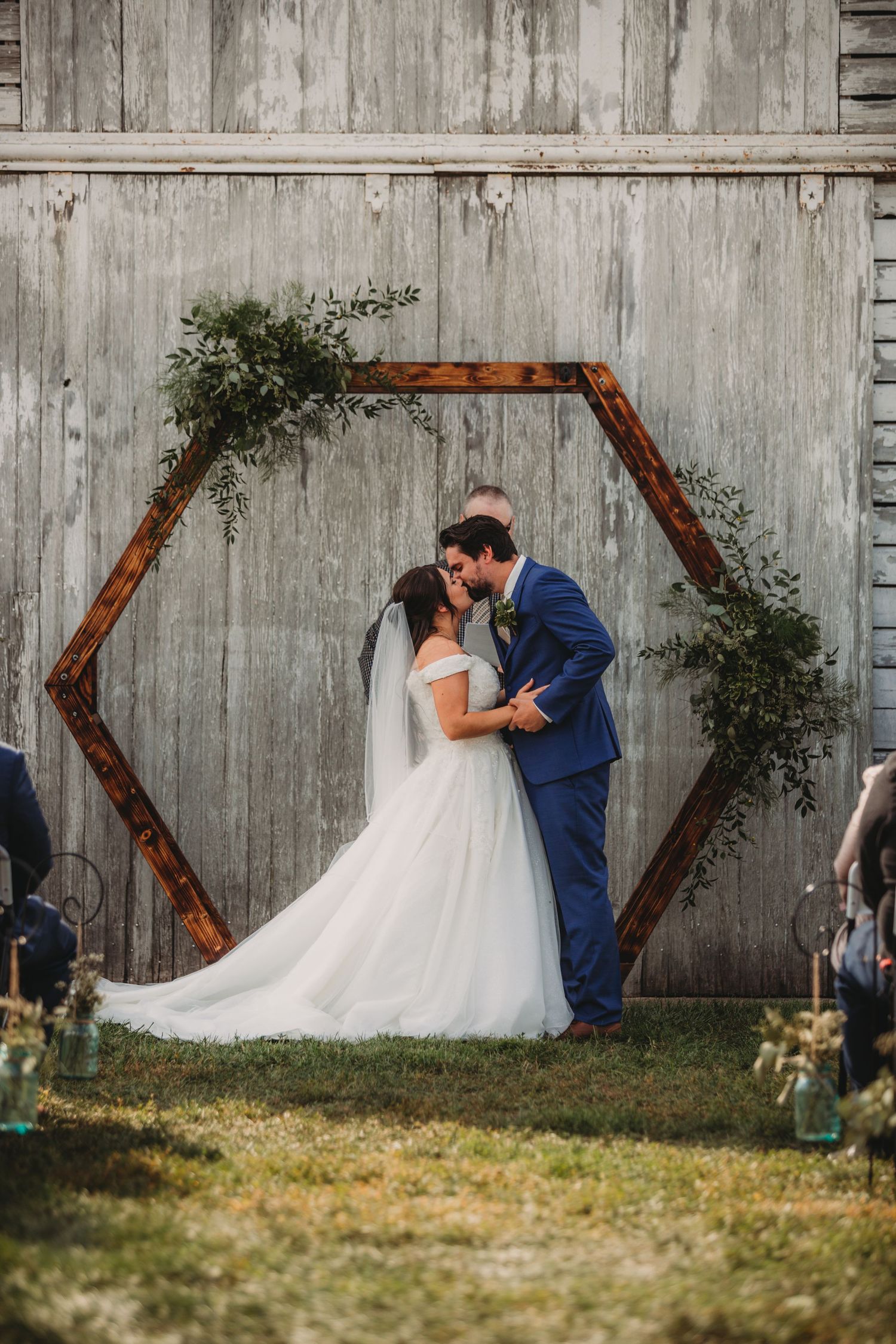 A rustic barn wedding ceremony featuring a wooden hexagon arch decorated with greenery.