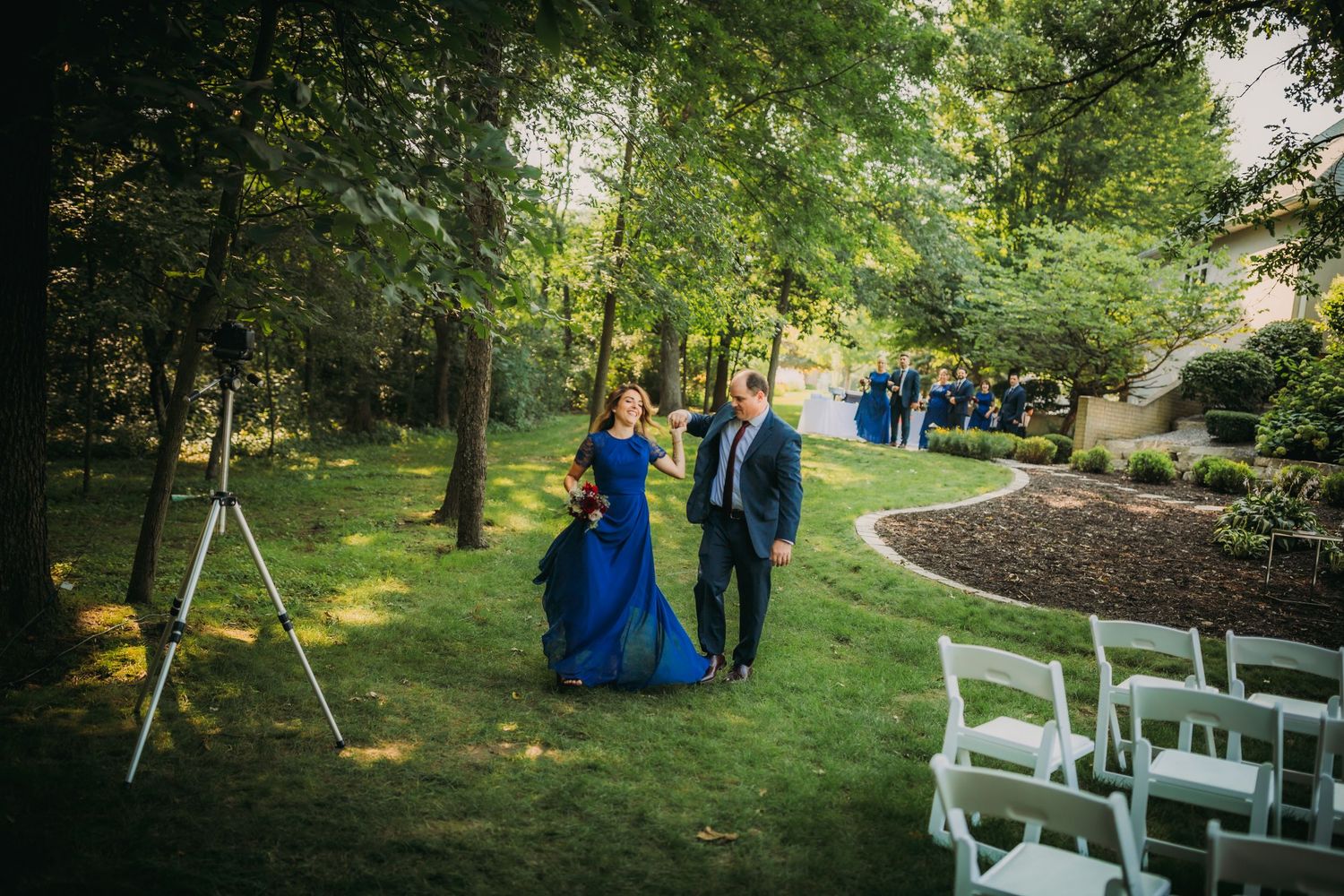 Garden wedding ceremony setup with white chairs and couple in blue walking path.
