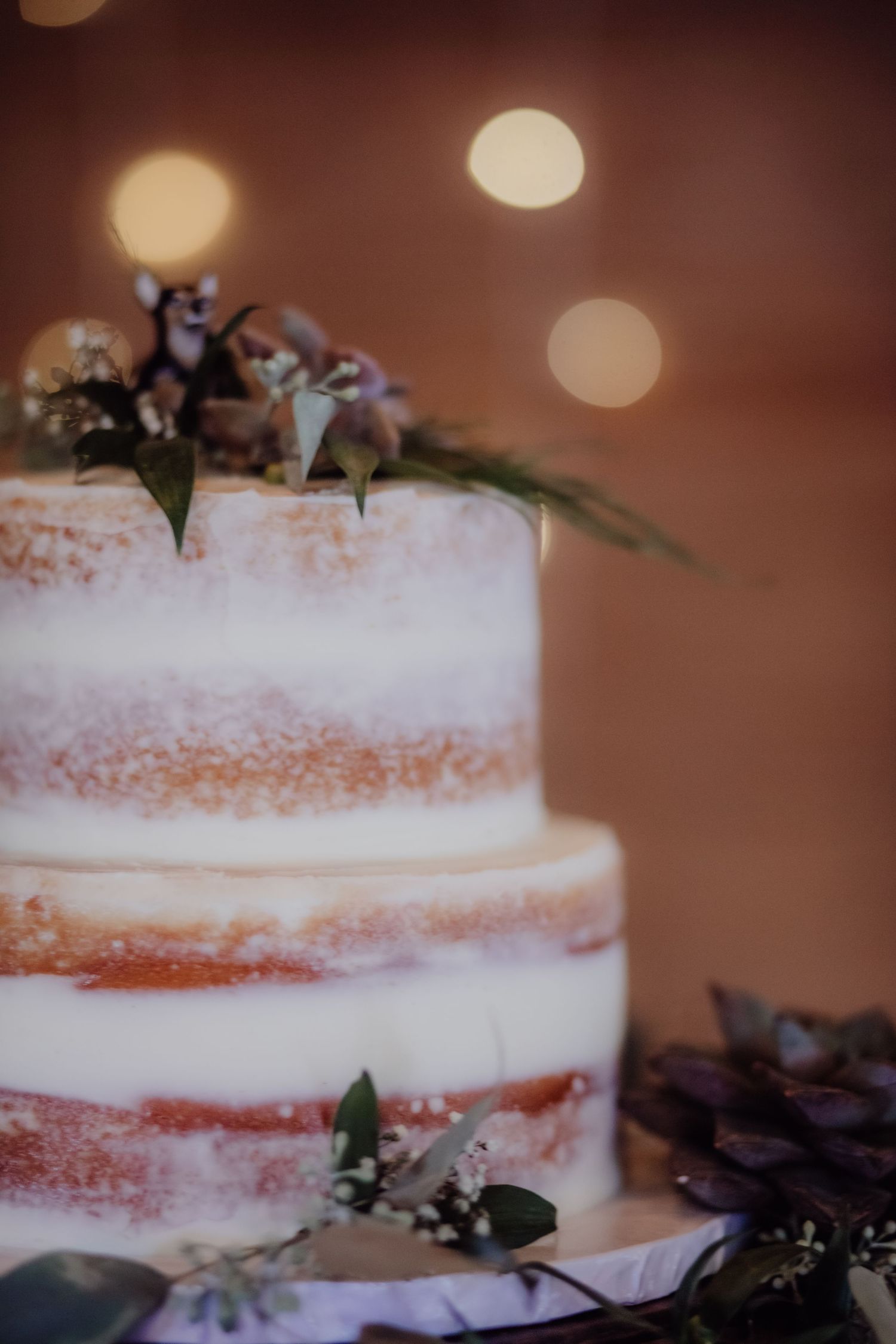 Elegant tiered wedding cake decorated with greenery against warm bokeh background.