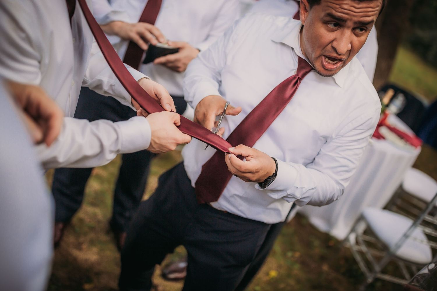 Groomsmen in white shirts and red ties examine details at an outdoor event.