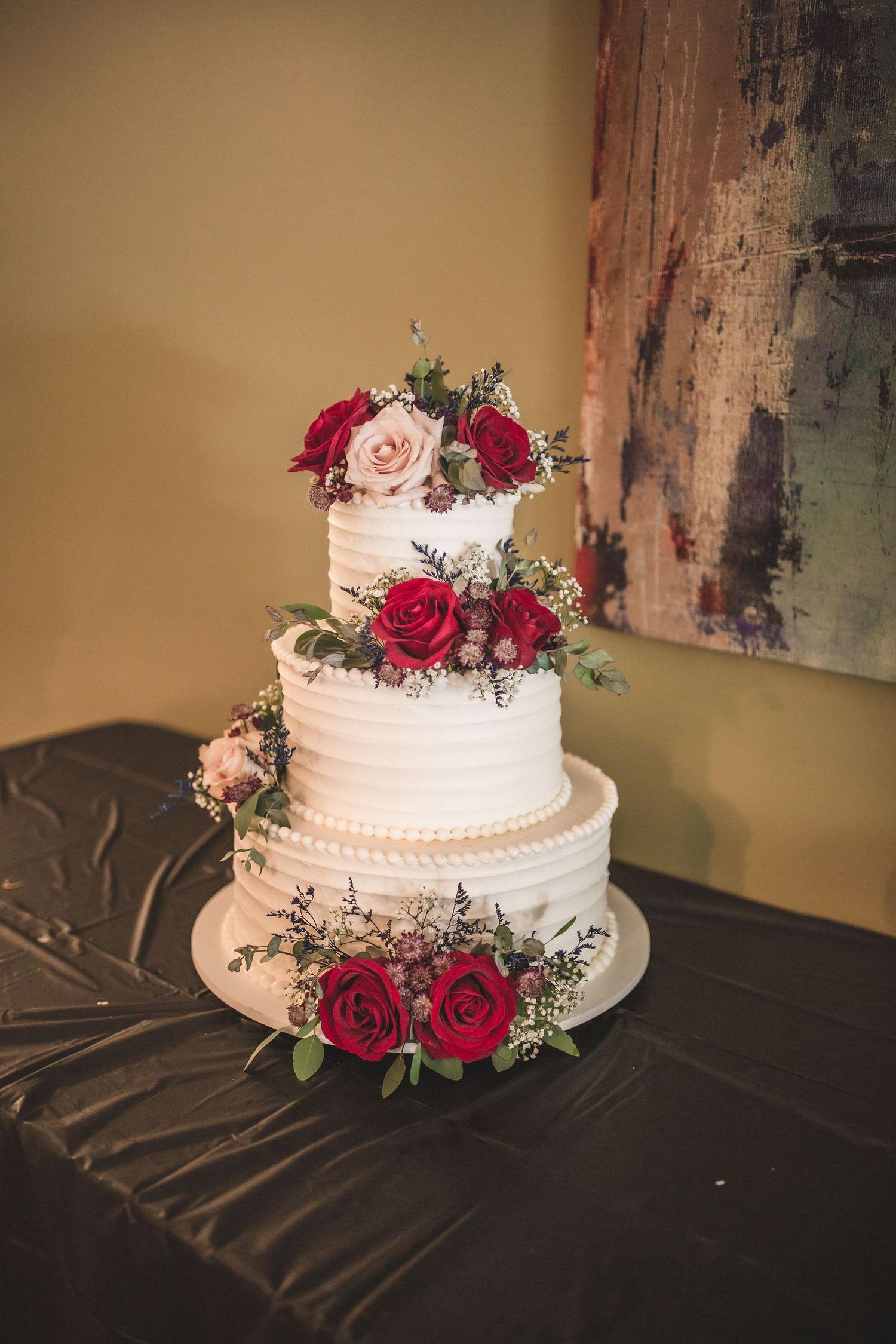 Close-up of a three-tiered white wedding cake adorned with red and blush roses against a rustic backdrop.