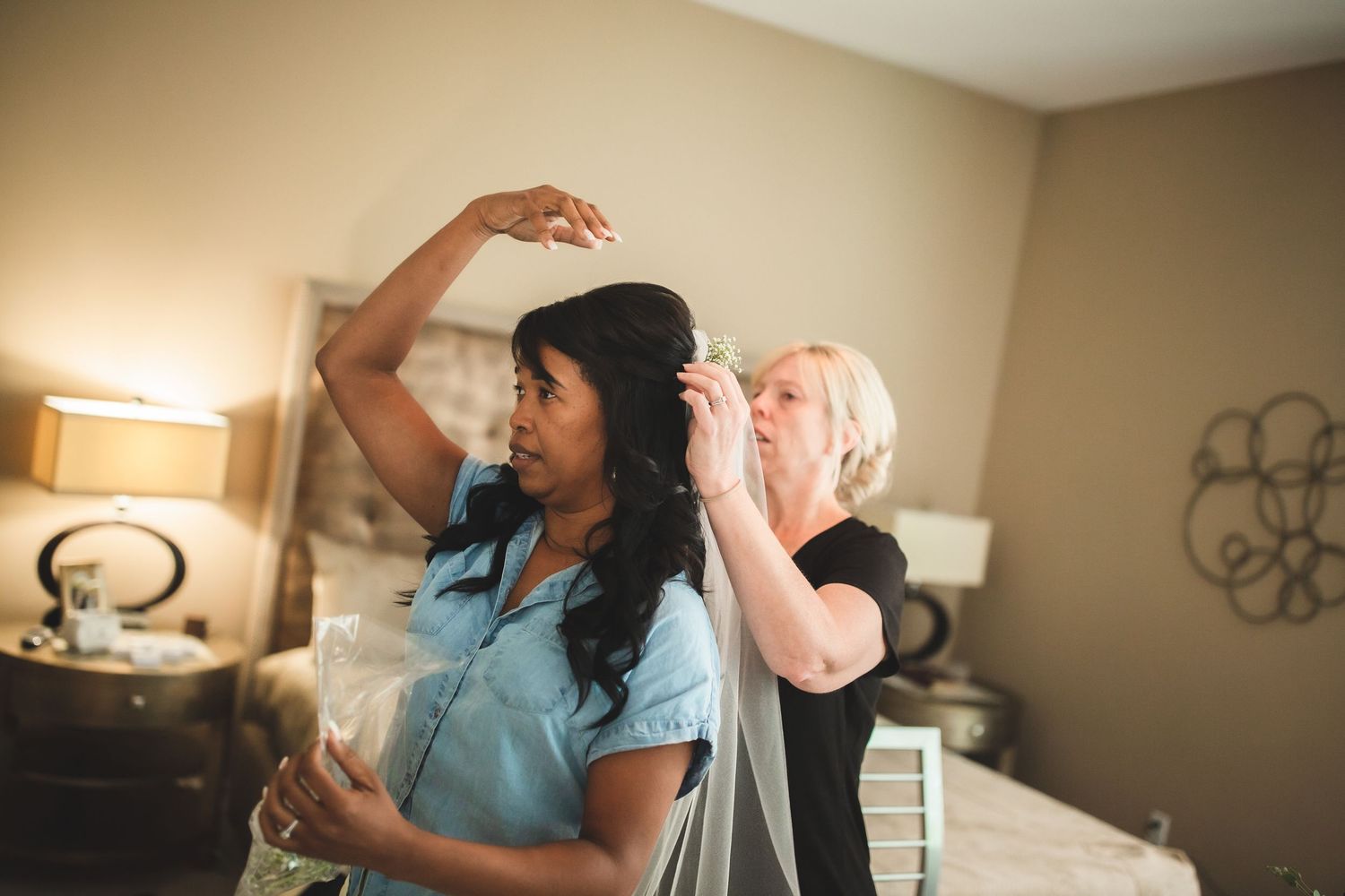 Wedding preparation scene showing stylist adjusting bride's veil in bedroom setting.
