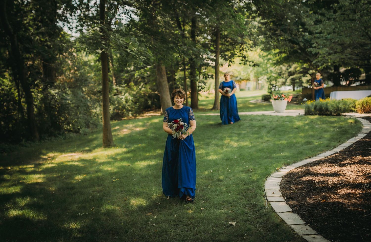 Bridesmaids in royal blue dresses walk along curved garden path holding bouquets.