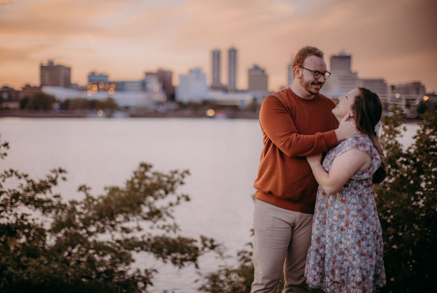 A couple embraces while standing on a riverside overlook with urban buildings in the warm evening light.