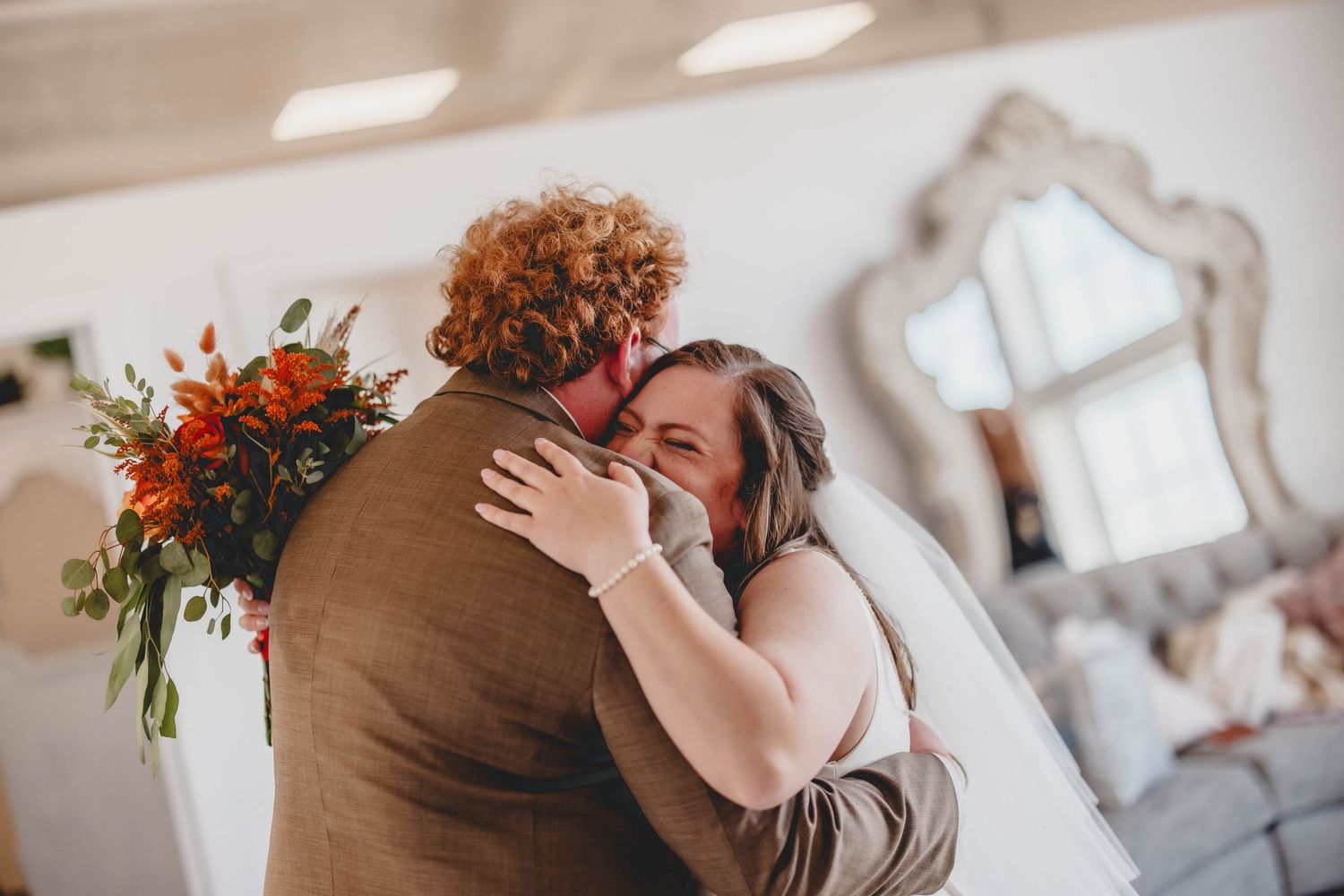 Romantic bridal couple embrace during first look in bright indoor setting with decorative mirror.