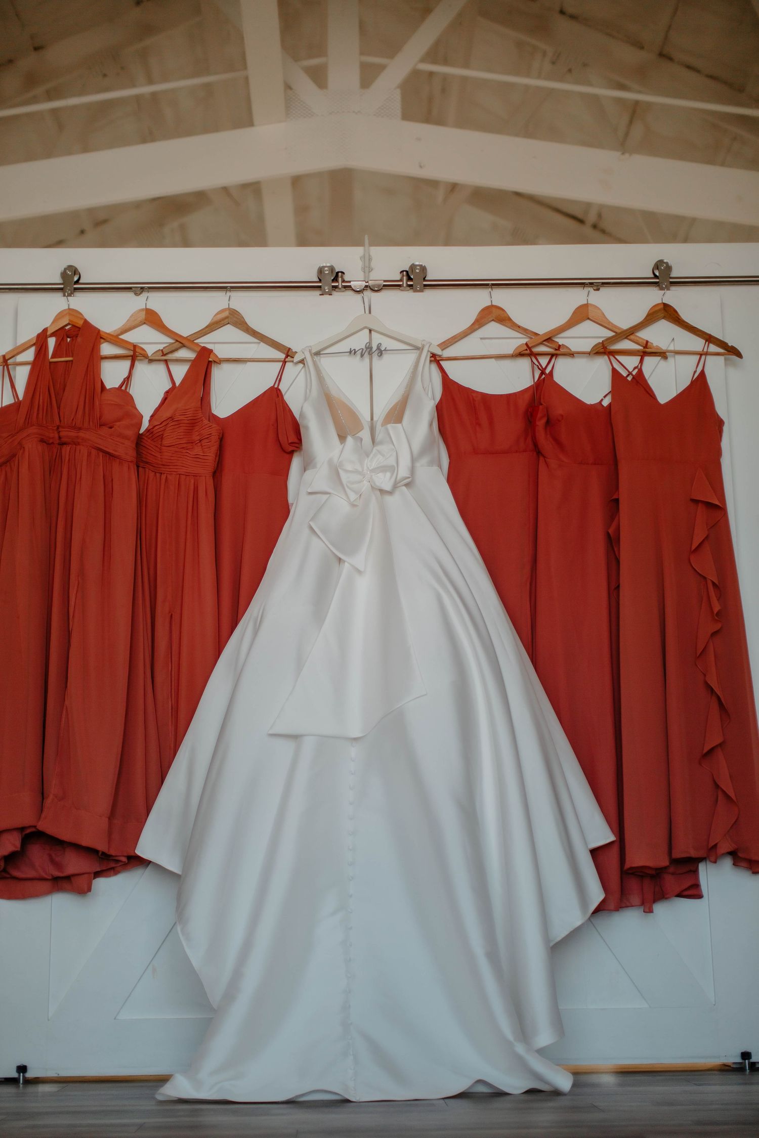 White satin wedding gown displayed between coral-colored bridesmaid dresses on wooden hangers.