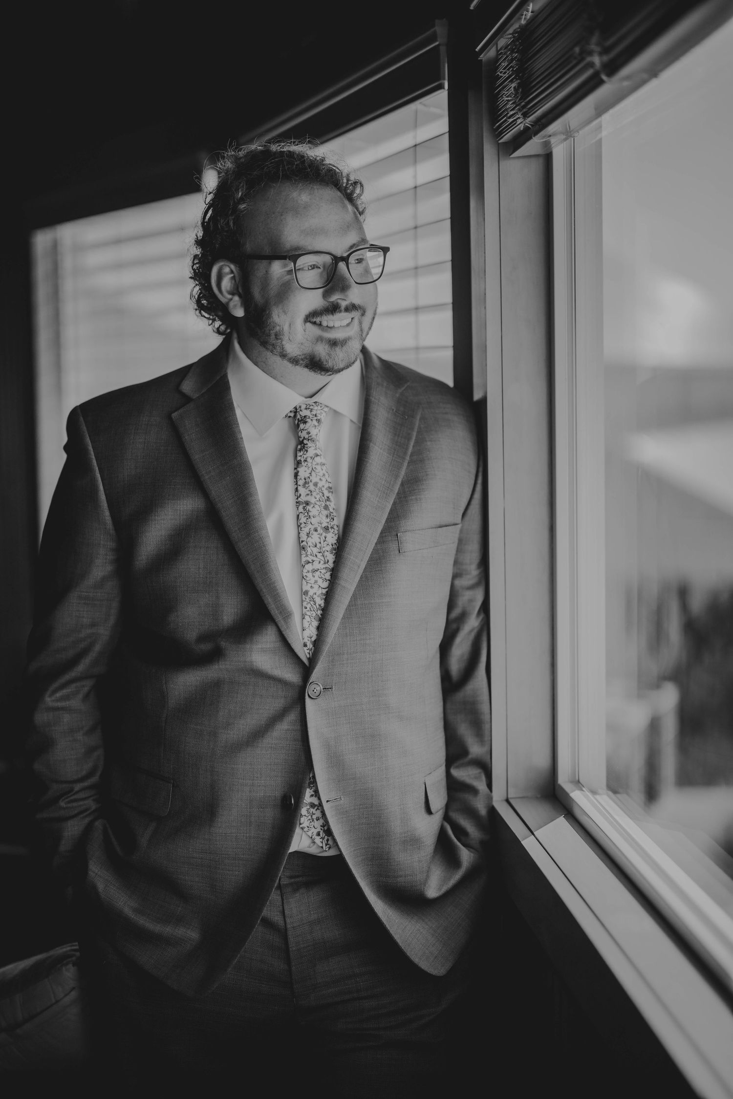 Black and white portrait of someone in business attire smiling while standing near a window.