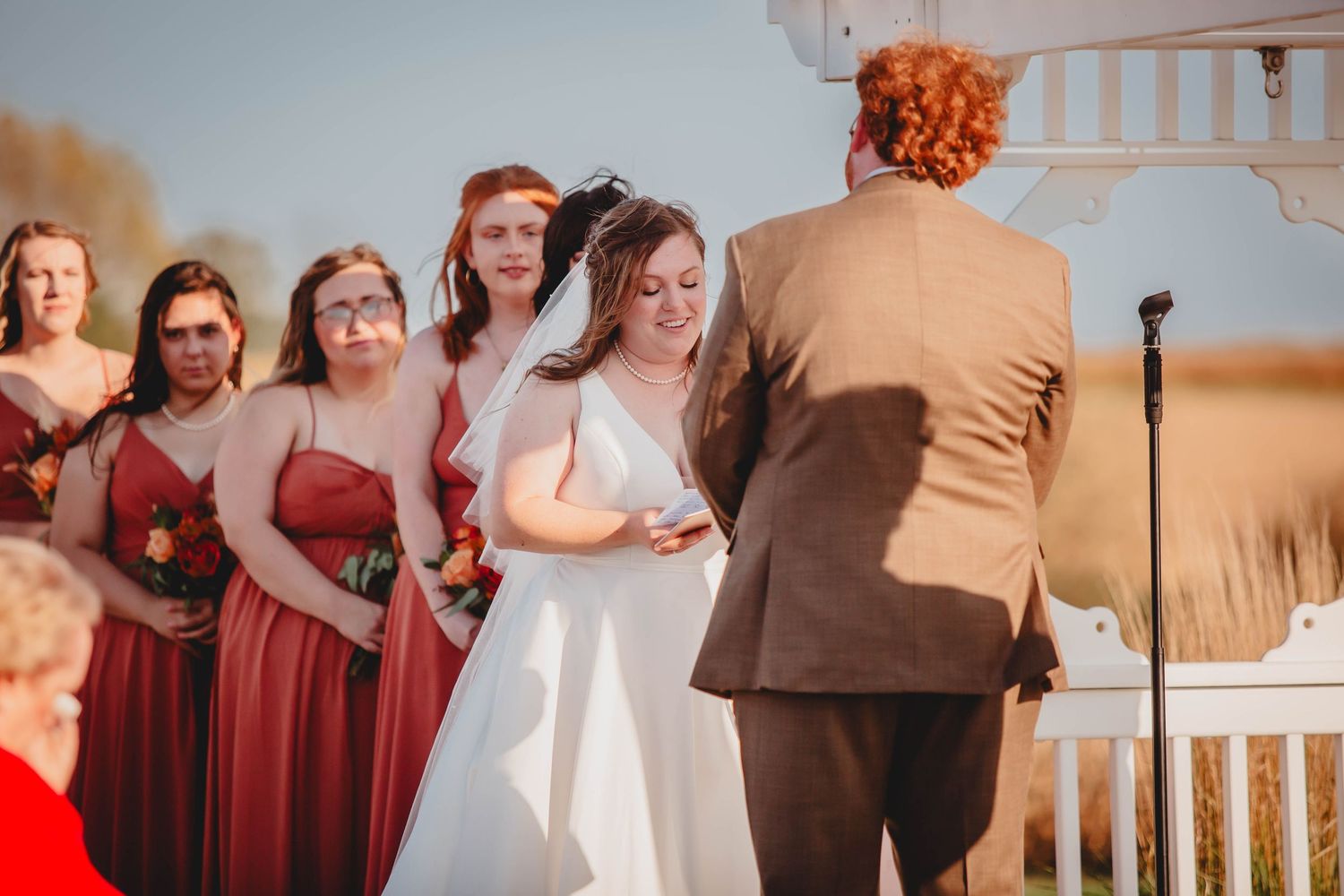 Outdoor wedding ceremony with bridal party in rust-colored dresses under white pergola.