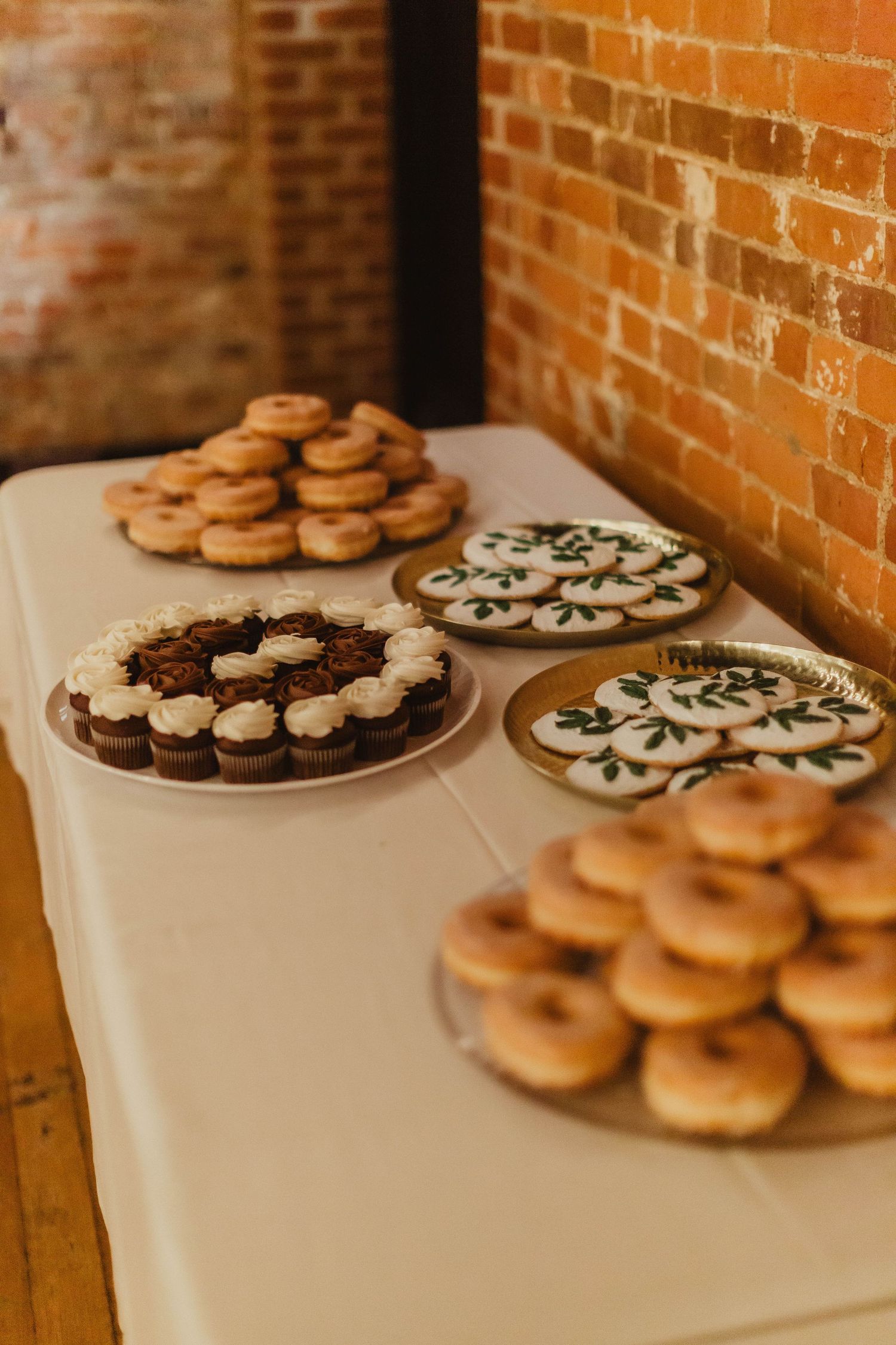 Assorted decorated cookies displayed on white tablecloth against exposed brick wall.