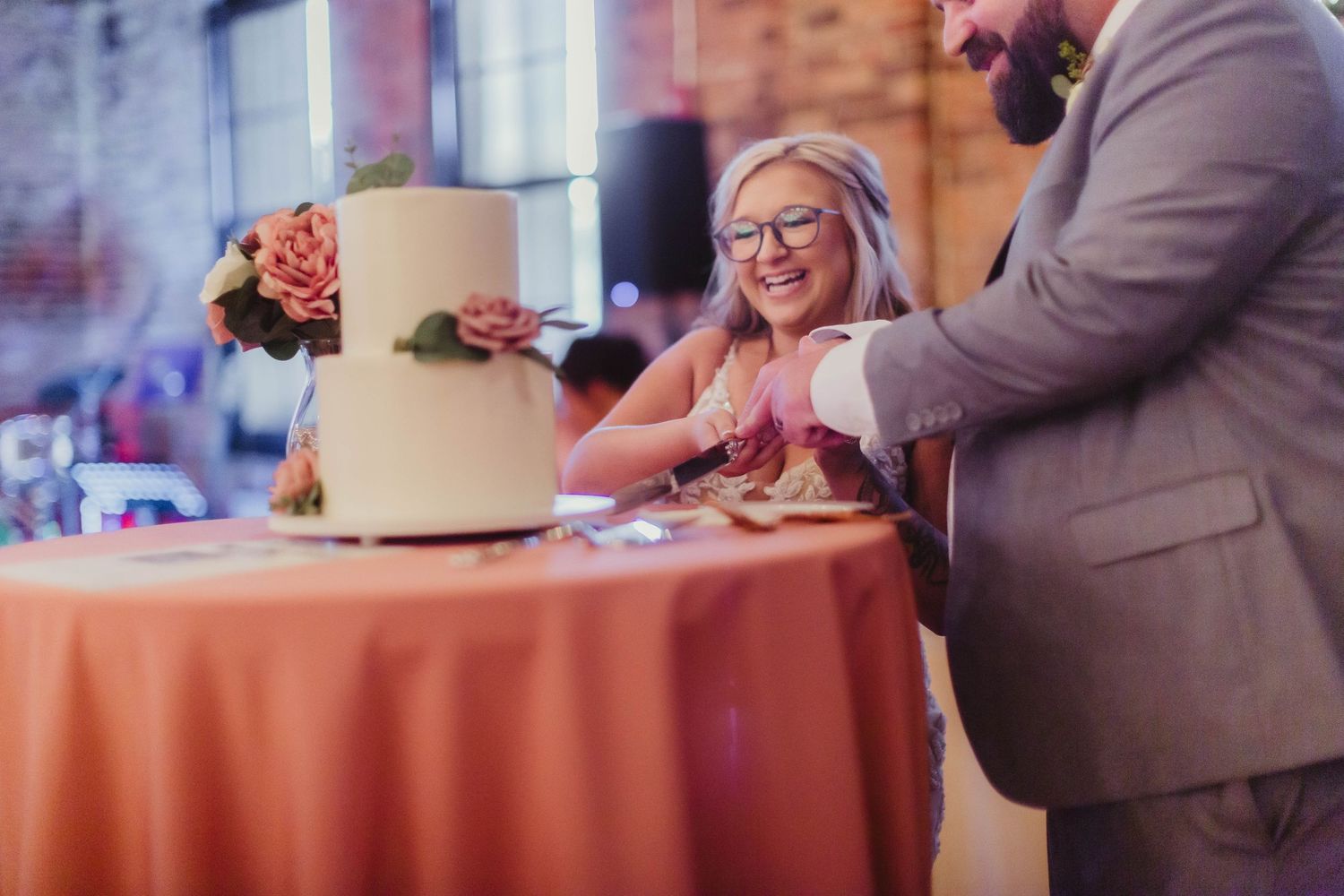 Couple cuts their simple two-tiered wedding cake decorated with coral flowers on a peach tablecloth.