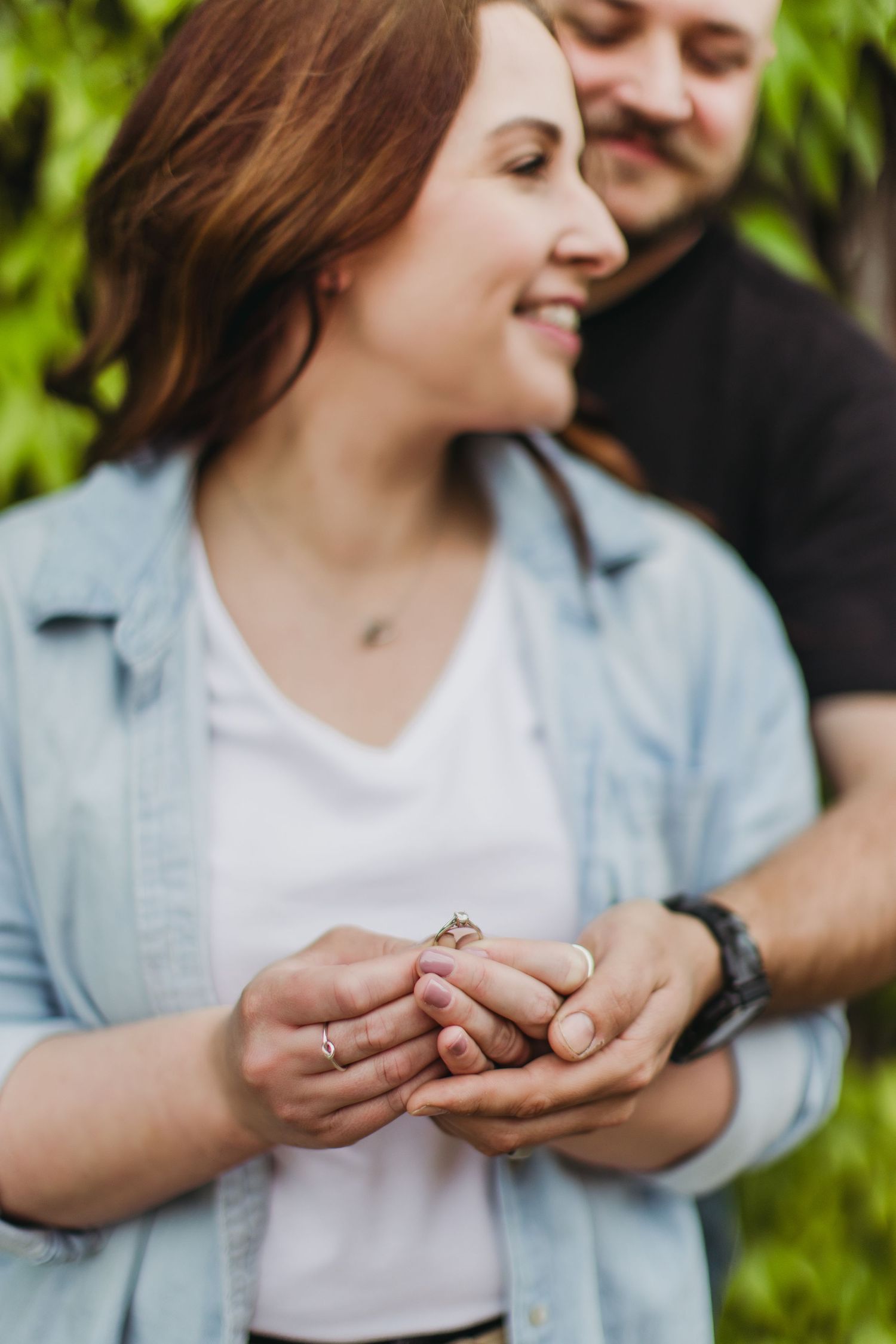 Happy couple sharing an intimate moment while looking at ring against green foliage.