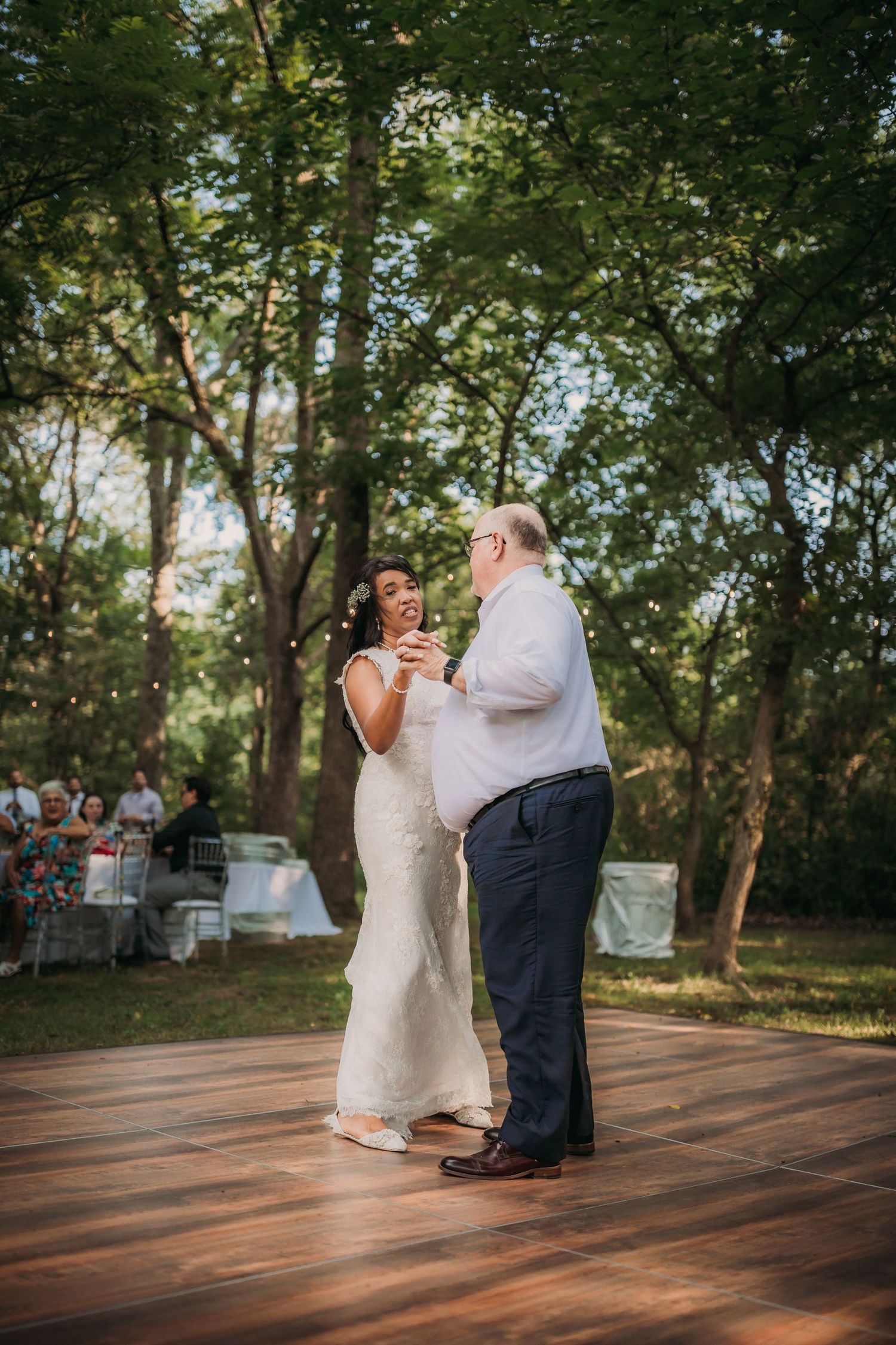 A couple dancing together on a rustic wooden deck with guests seated in the background at an outdoor reception.