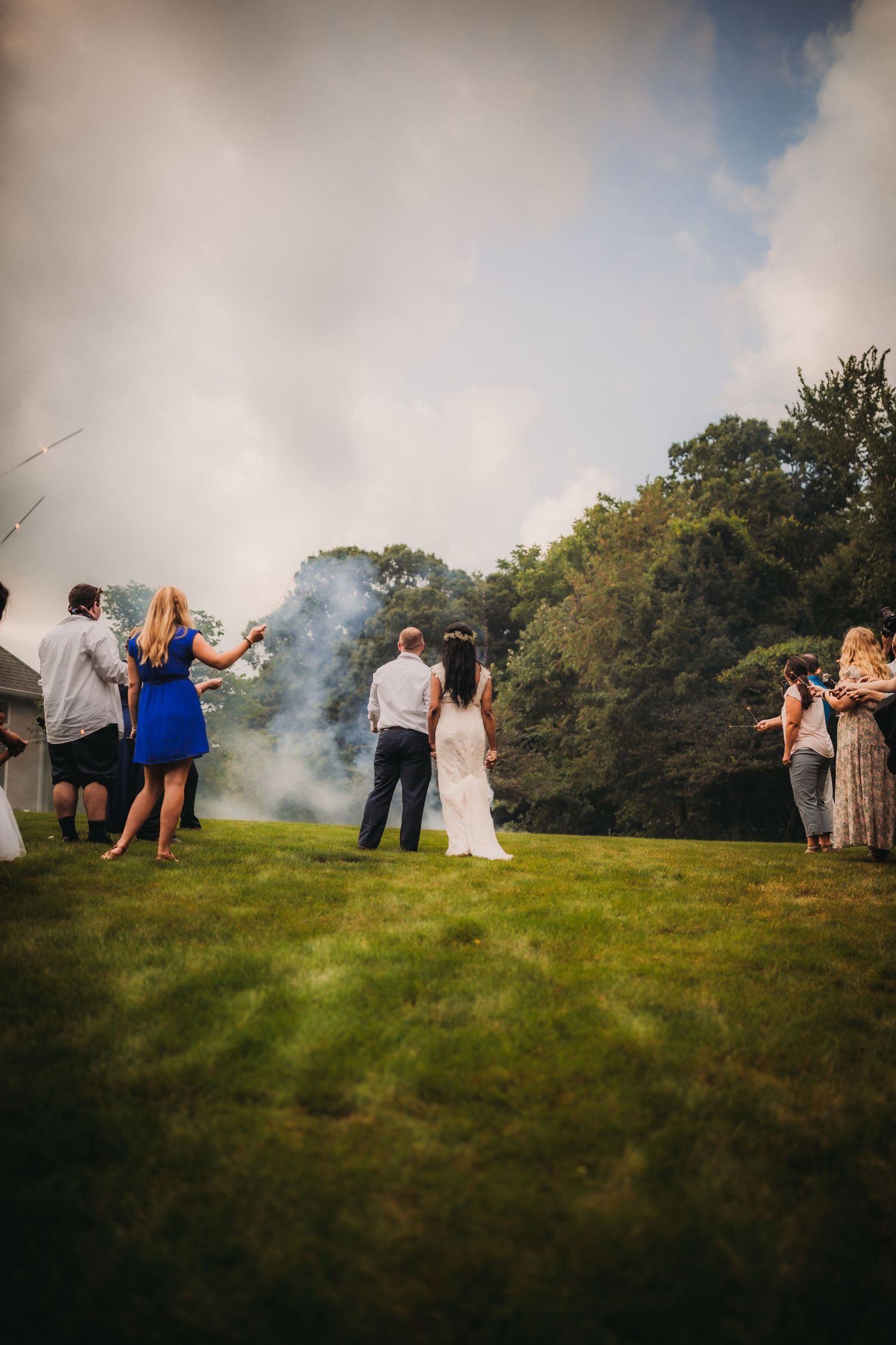 Crowd gathers on a grassy knoll for an outdoor wedding celebration with dramatic smoke effects.