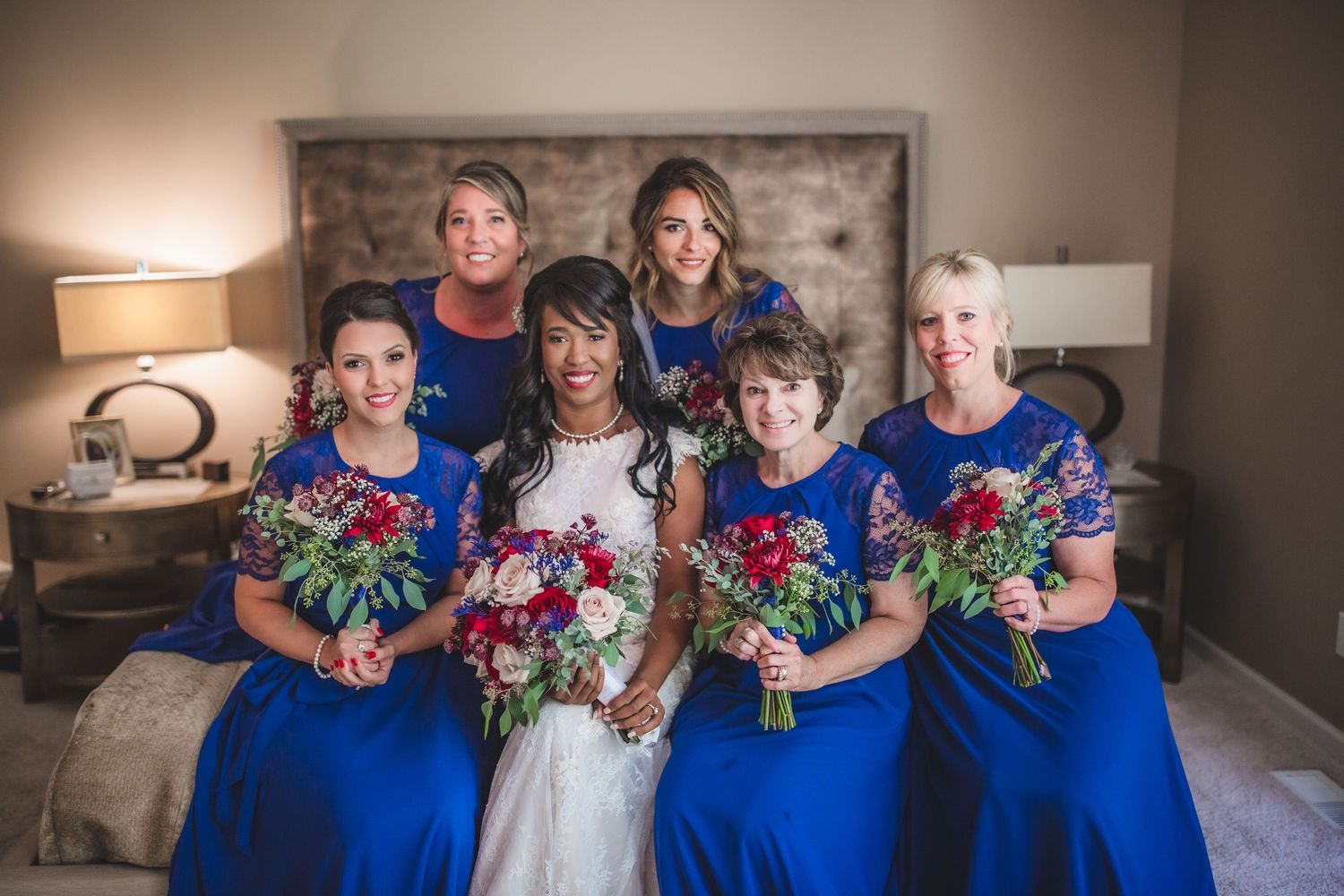 Wedding party in blue dresses and white gown hold colorful bouquets in bedroom setting.