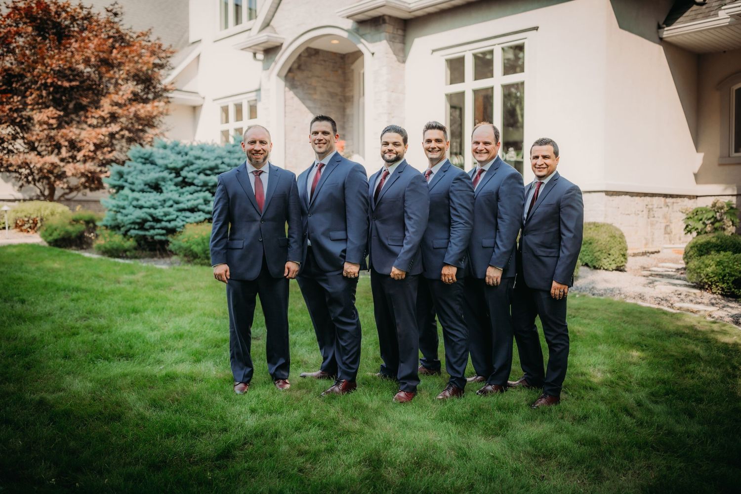 Wedding party members posed in navy blue suits and ties on a manicured lawn.
