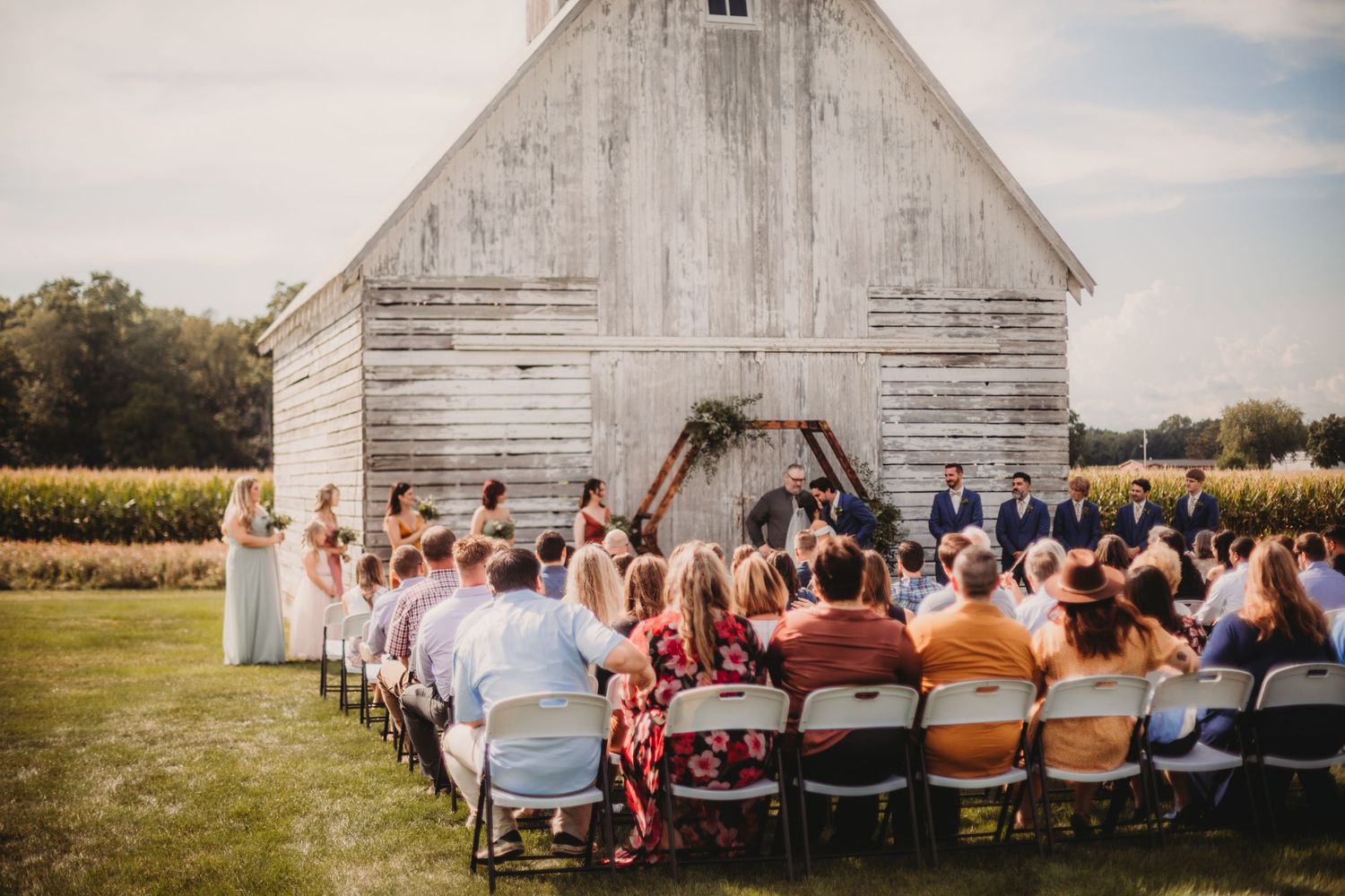 Rustic barn wedding ceremony setup with white chairs arranged in rows on grass under blue sky.