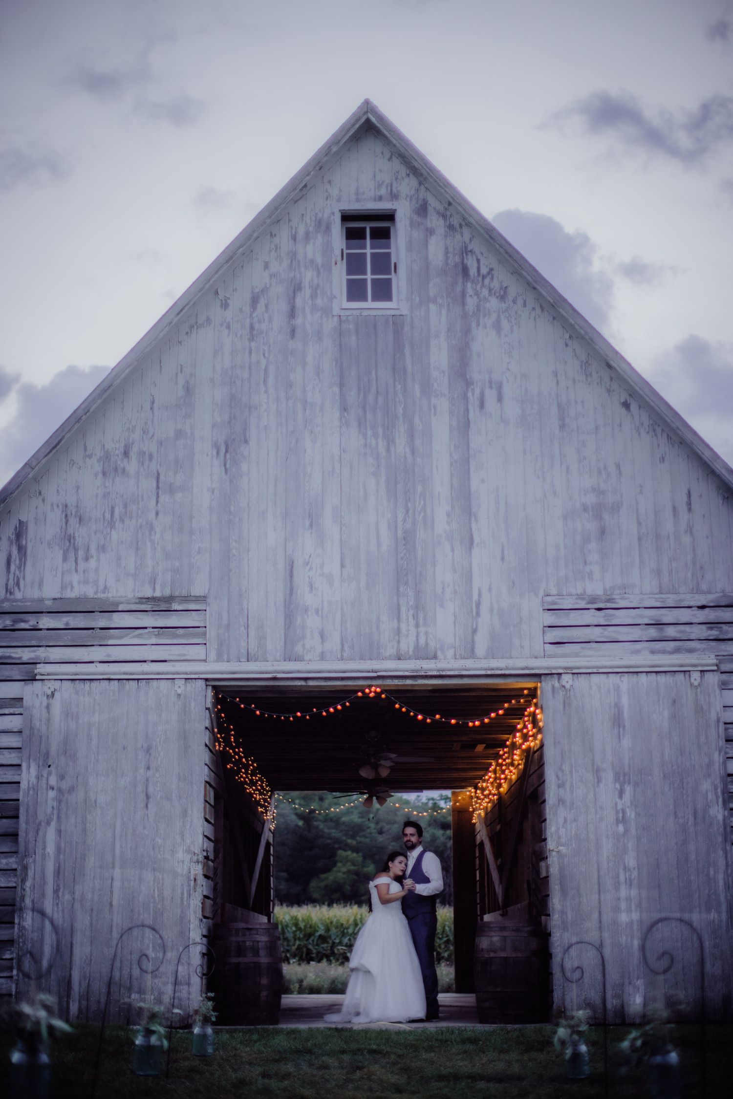 A rustic white barn with string lights frames a romantic wedding scene in the doorway at dusk.