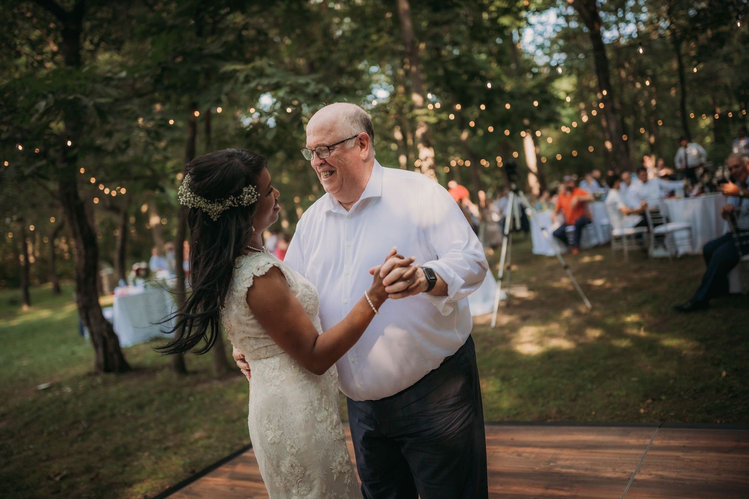 Couples share a dance on wooden platform during outdoor reception with string lights and forest backdrop.