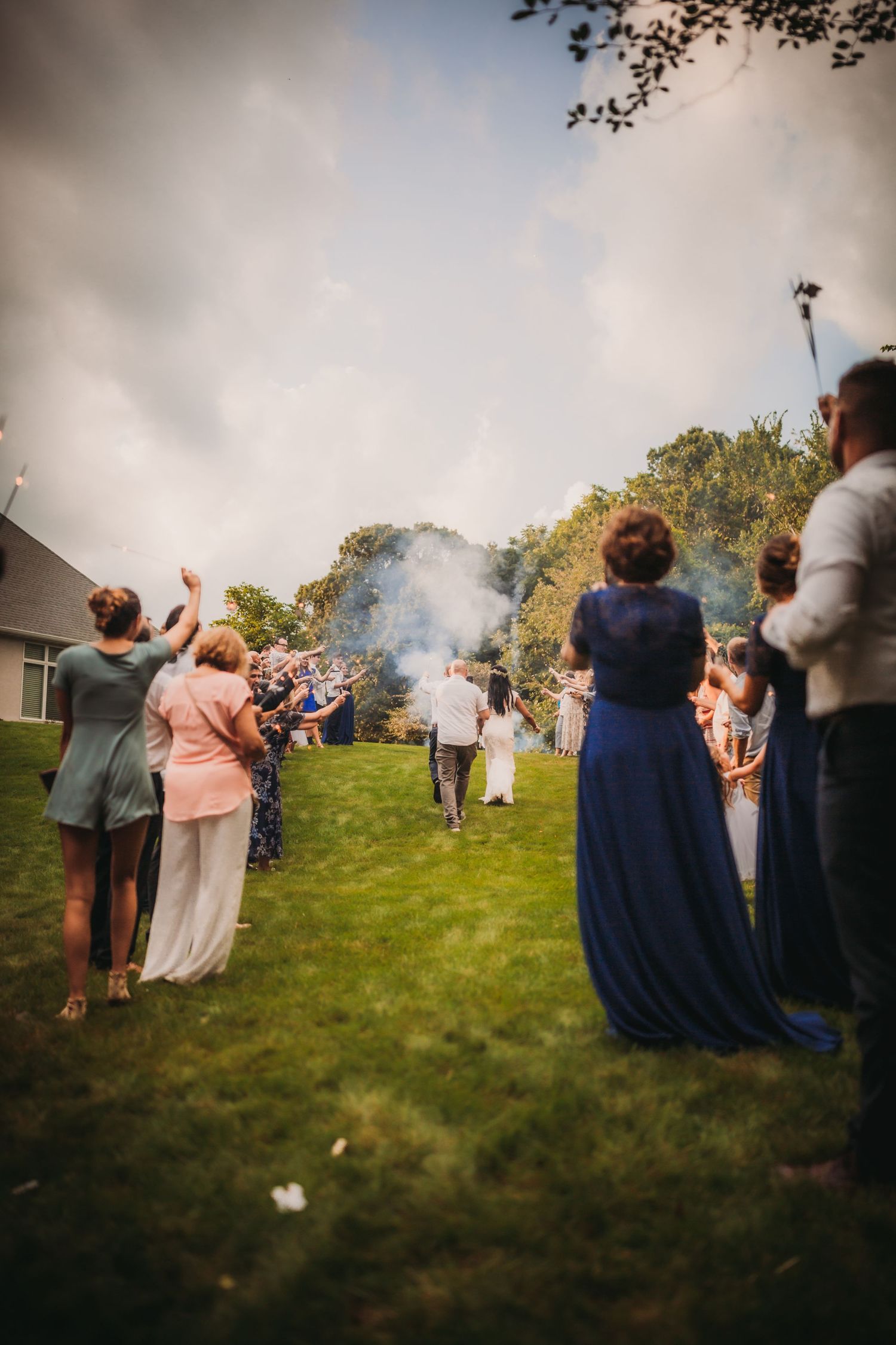 Wedding party and guests stand on a green hill during a late afternoon outdoor celebration.