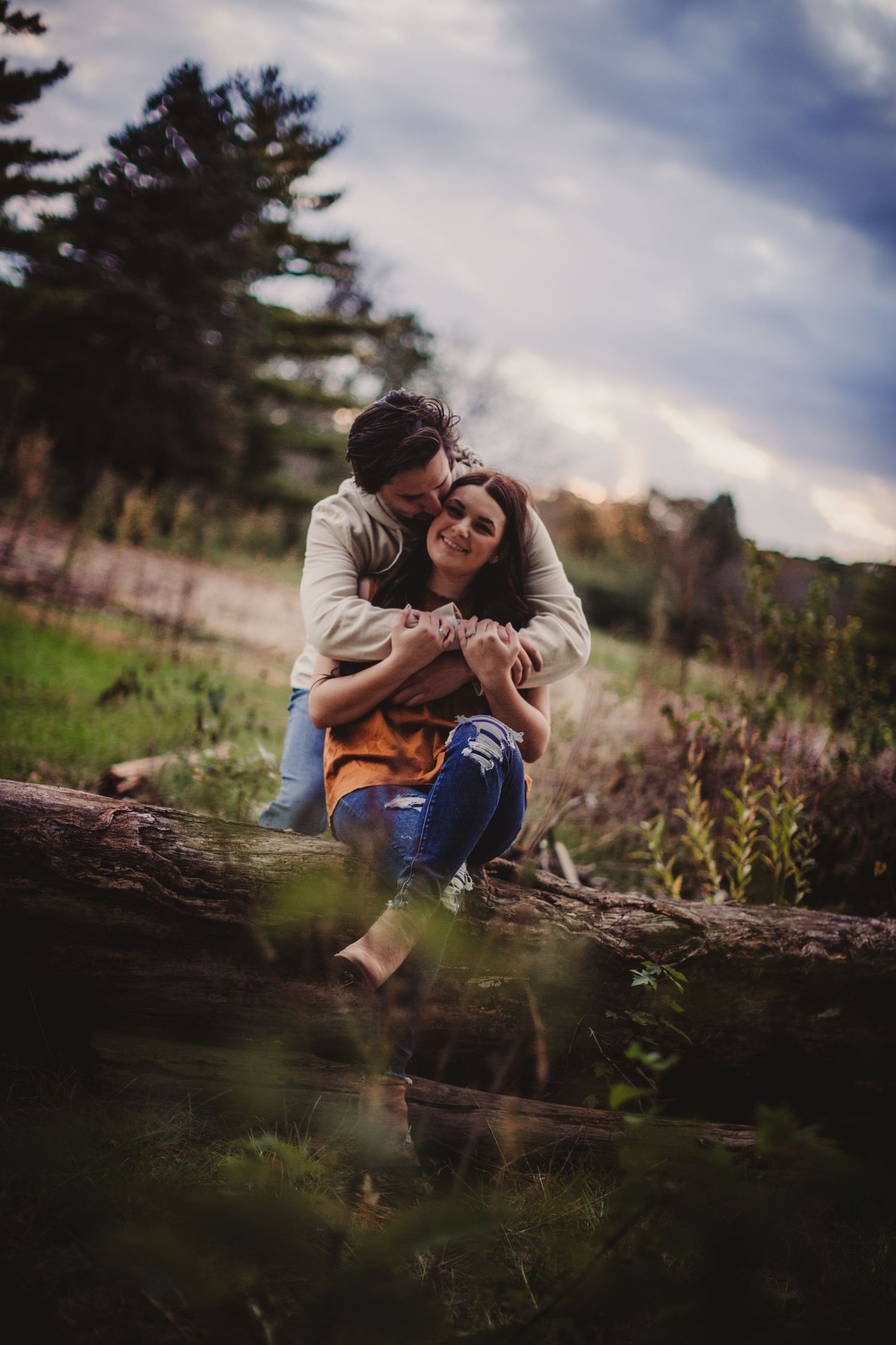 Couple embracing on wooden log with pine trees and cloudy sky in background.