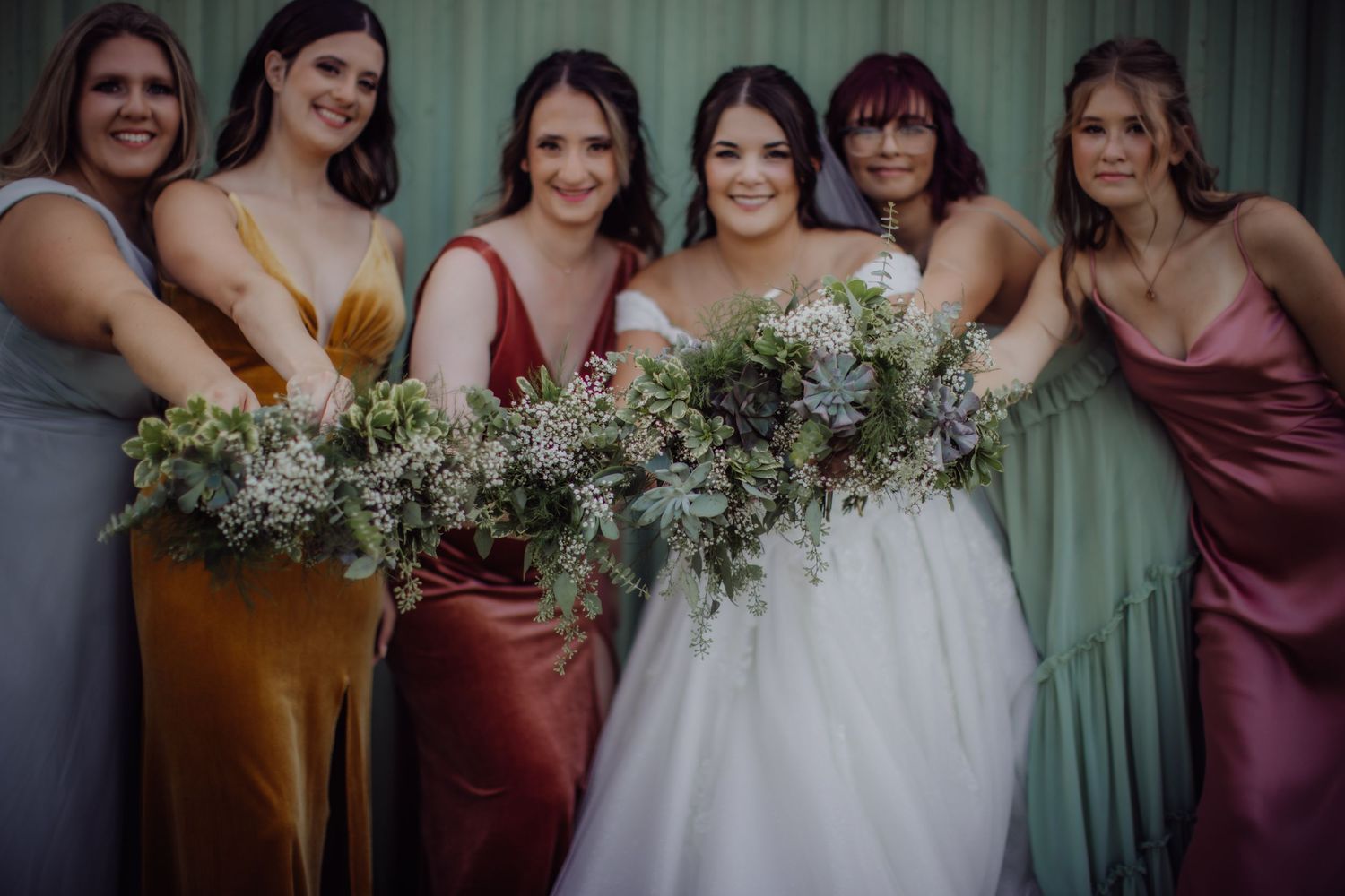 Bridesmaids in varied colorful dresses hold bouquets while posing together.