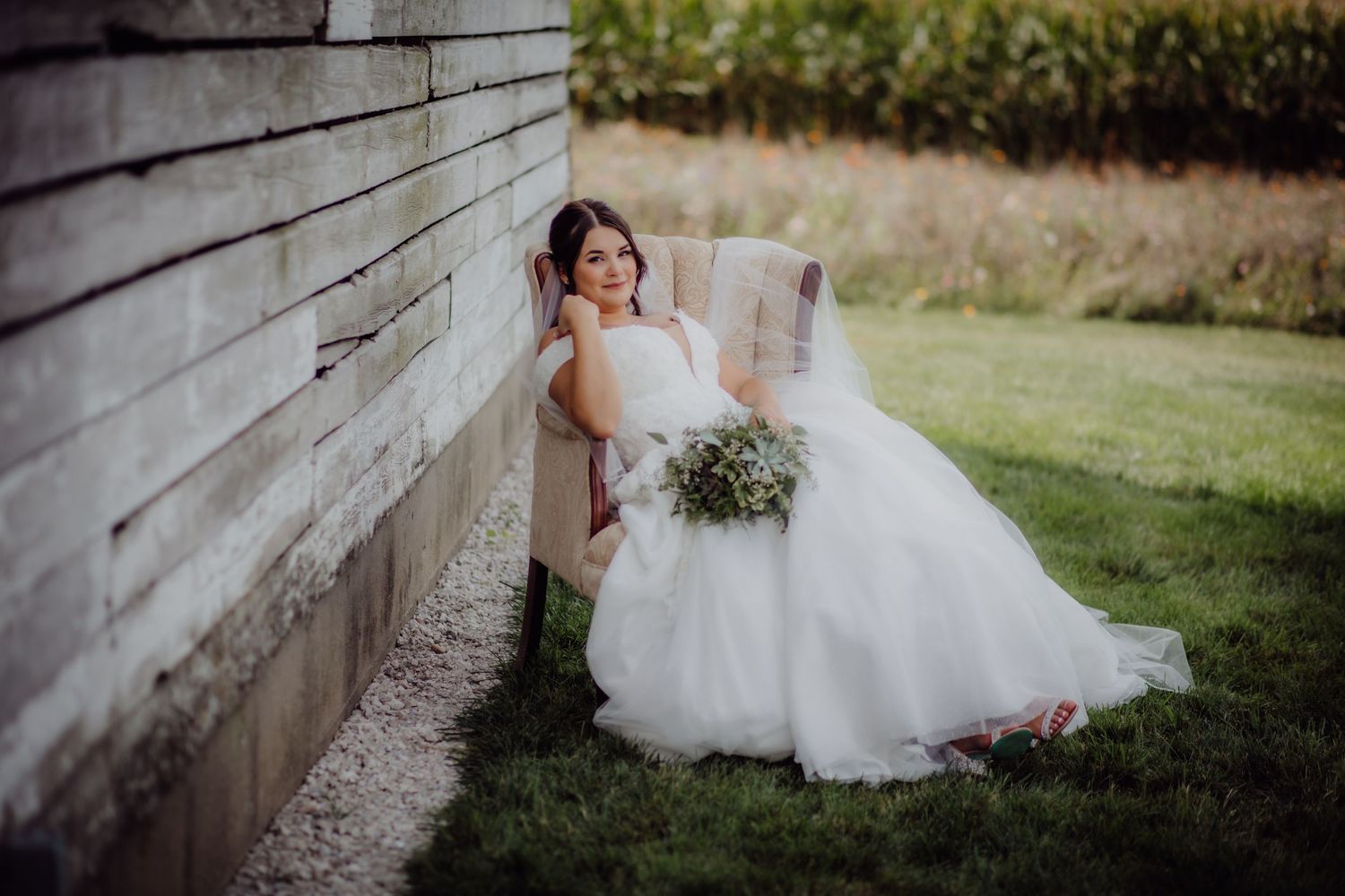 Elegant bride in white gown sitting beside weathered barn wall holding fresh floral bouquet.