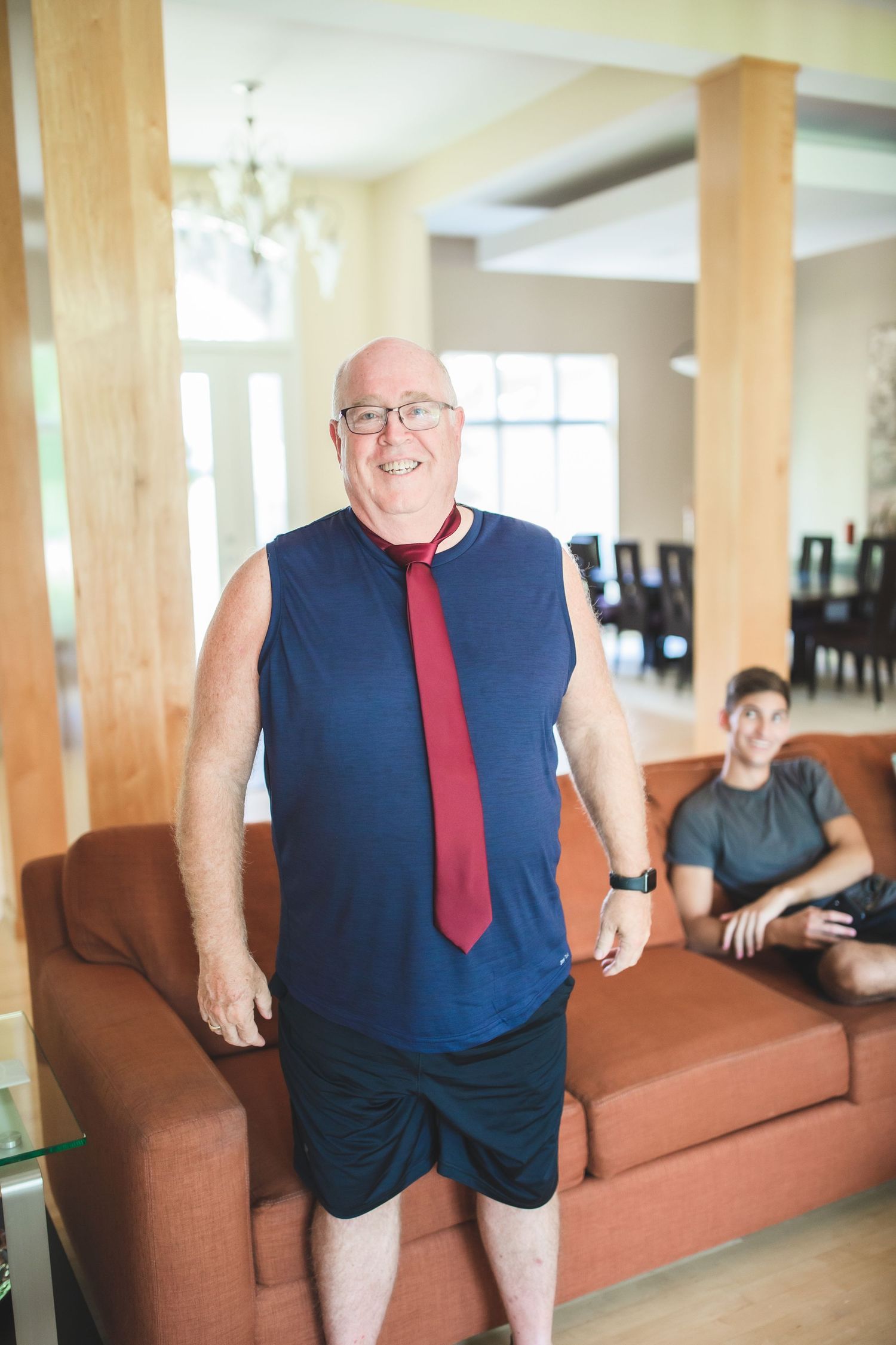 Person wearing a navy sleeveless top and red tie stands in front of a brown couch.