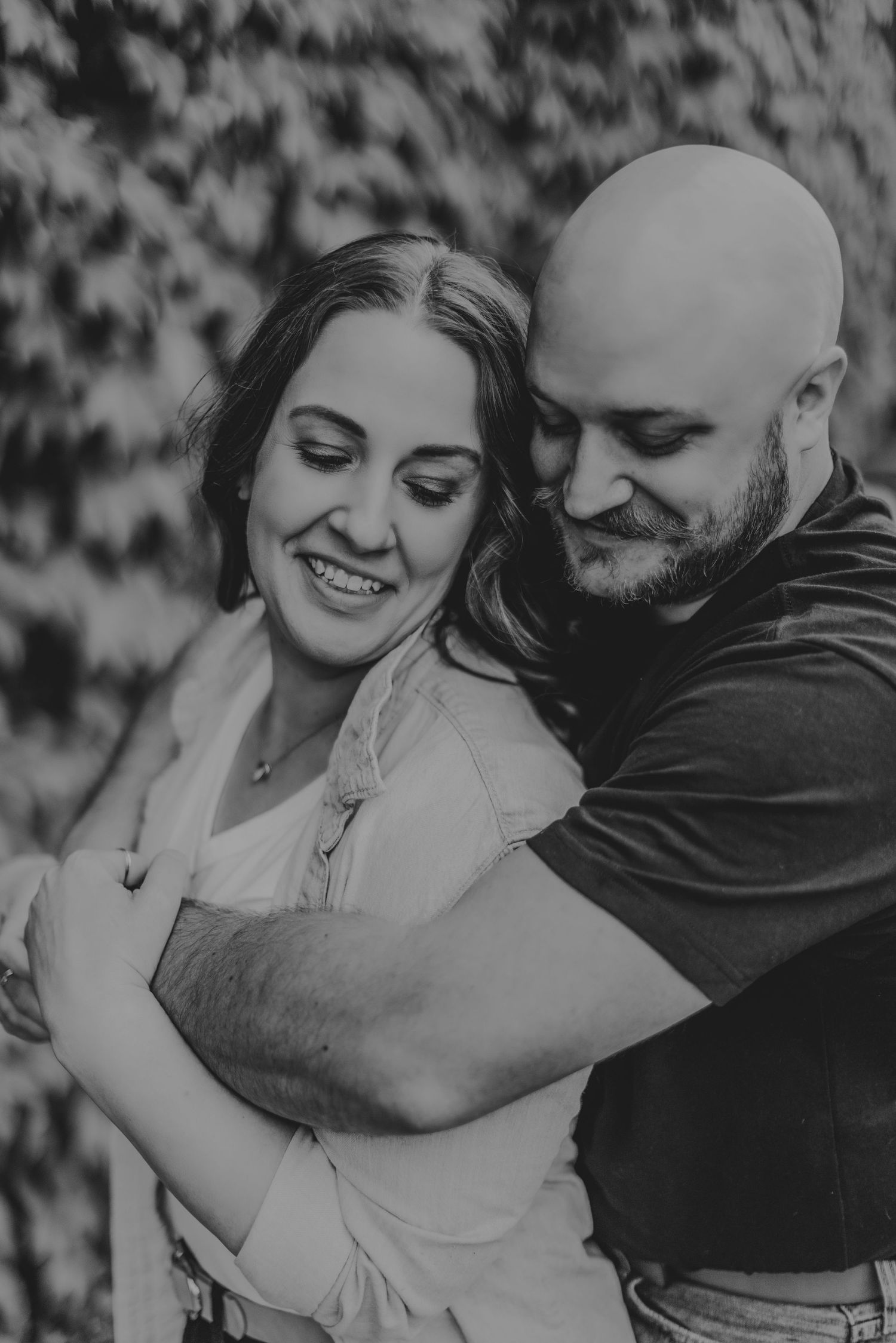 Black and white portrait of a couple sharing a loving embrace against an ivy-covered wall.