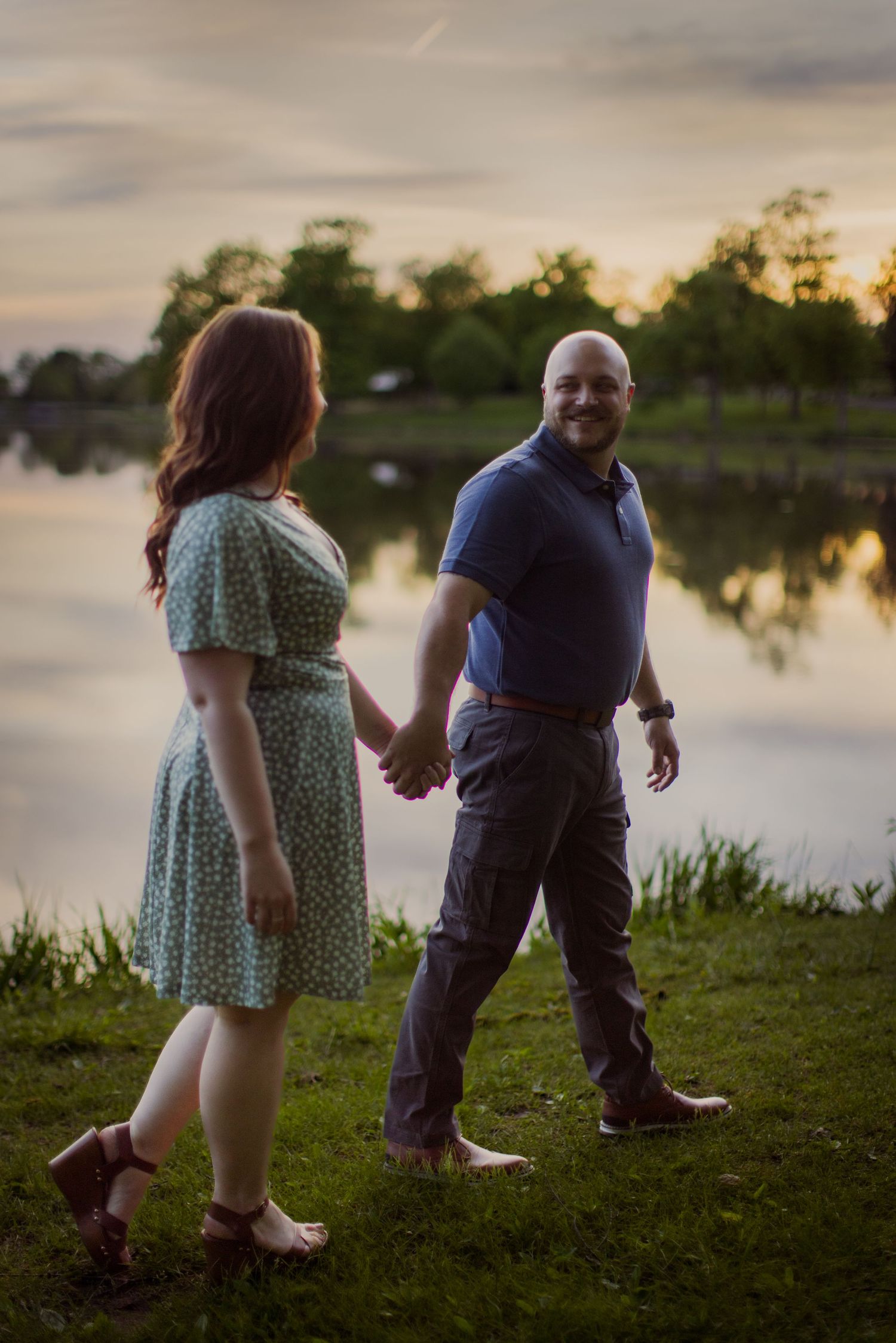 A couple walks along grassy lakeside path during sunset wearing casual attire.