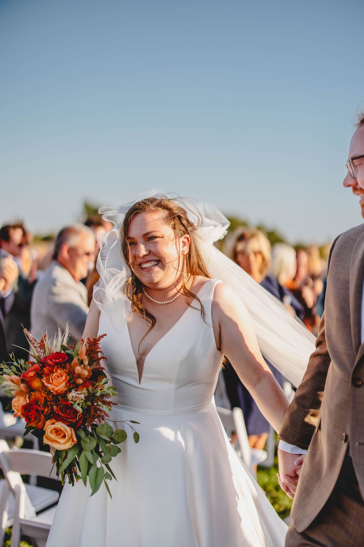 Bride holds fall-colored bouquet while laughing during outdoor ceremony exit.