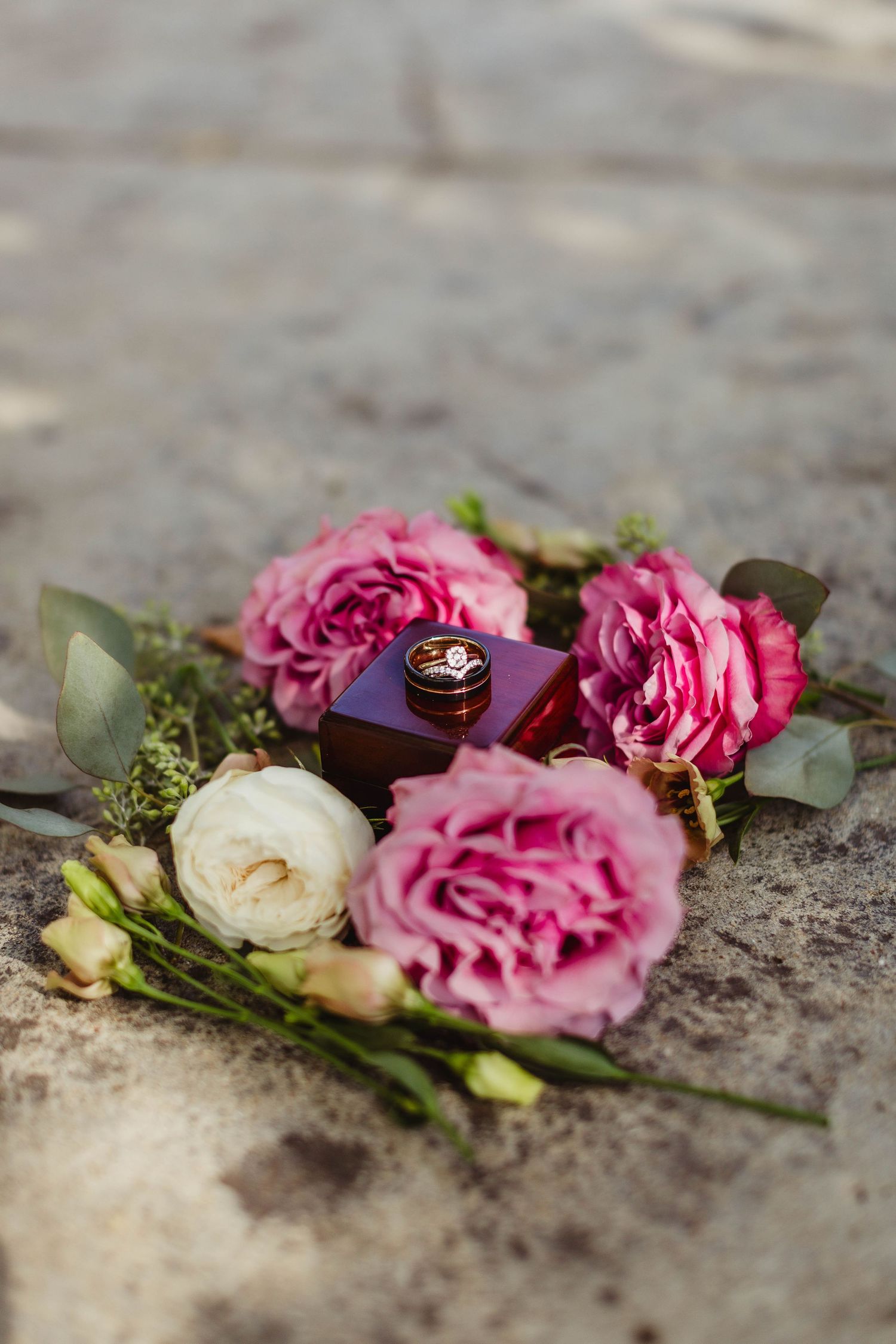 Wedding rings on wooden box surrounded by pink and white roses on stone surface.