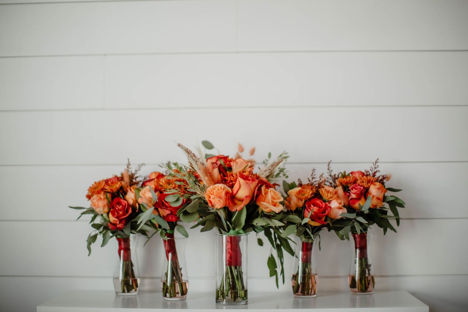 Row of five coral and orange floral bouquets arranged in glass vases against white shiplap wall.