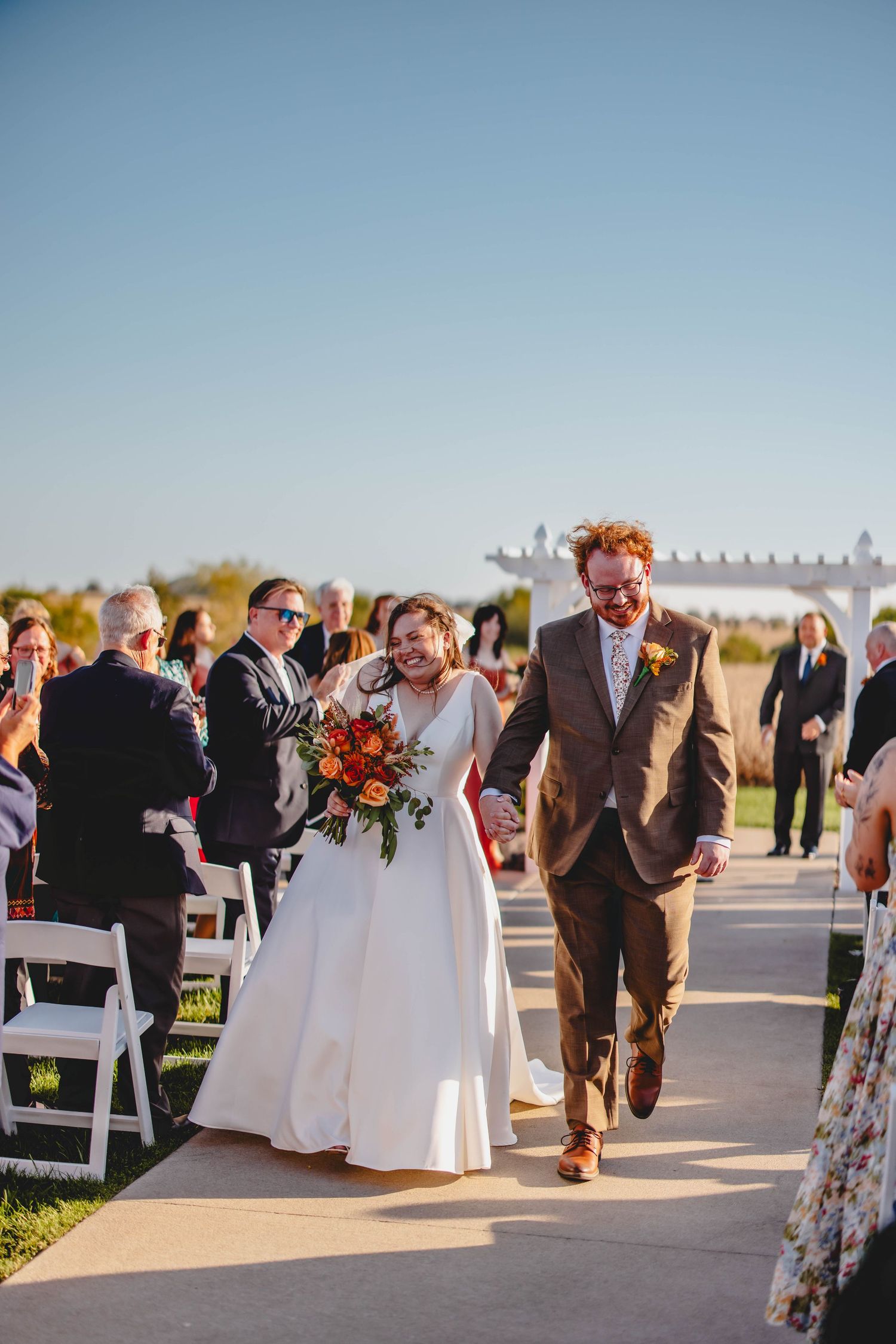 Joyful couple exits their outdoor wedding ceremony carrying vibrant fall bouquet.