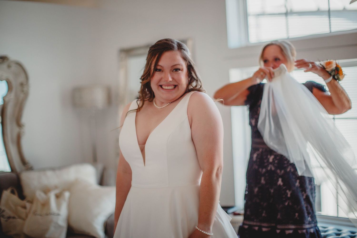 A bride in a classic white v-neck wedding dress smiles while getting ready in a bright room.
