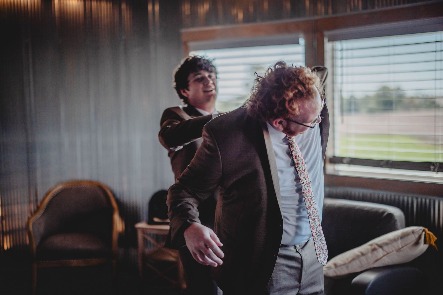 Two people in formalwear helping each other prepare in a room with windows.