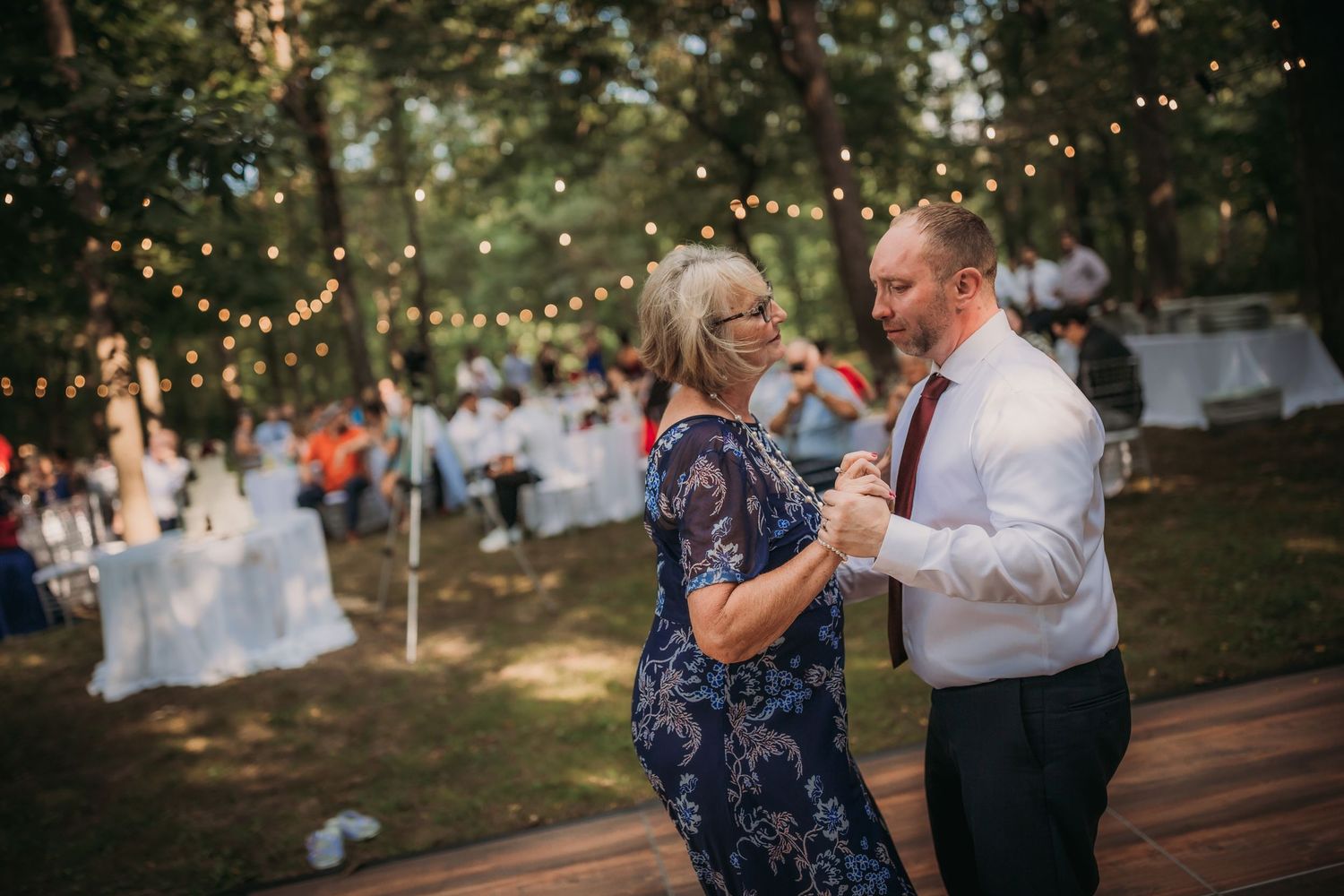 Dancing couple enjoys a special moment at outdoor woodland reception with fairy lights and guest tables visible.