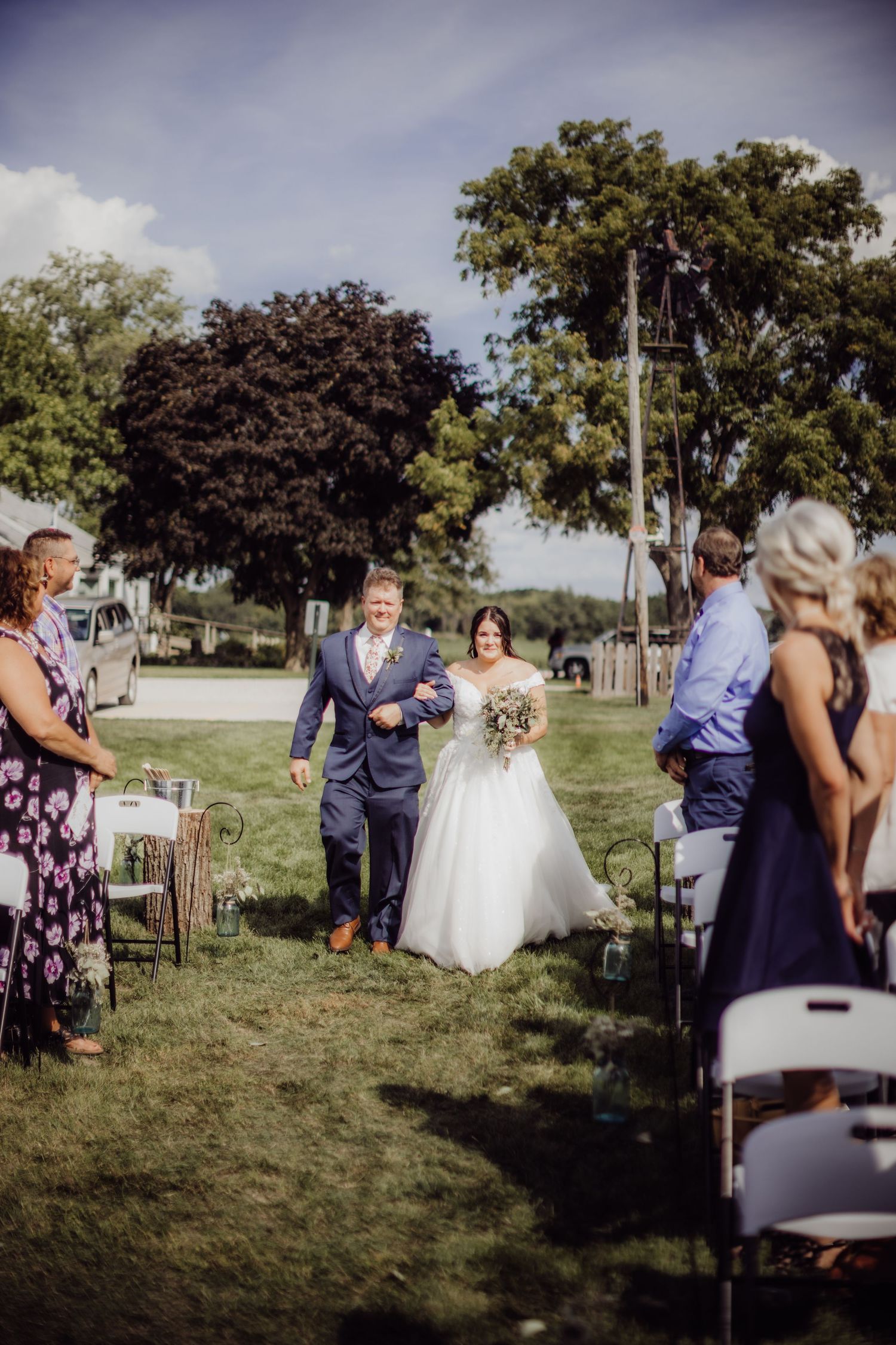 Wedding ceremony takes place outdoors with guests seated on white chairs amidst a natural landscape.