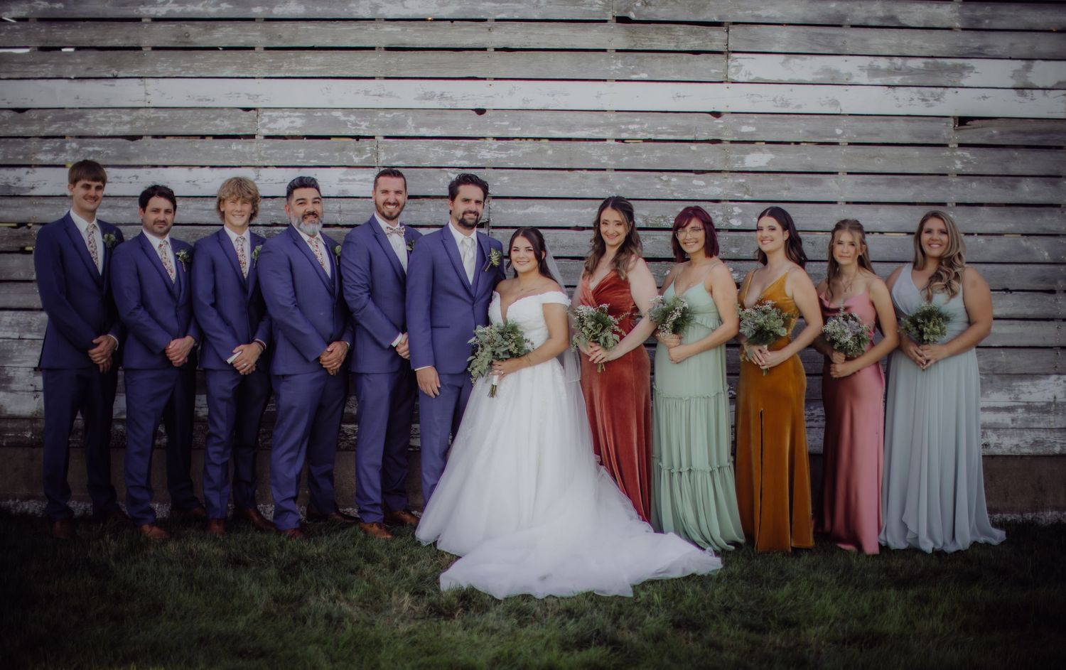Wedding party poses for formal group photos in front of a rustic wooden barn backdrop.