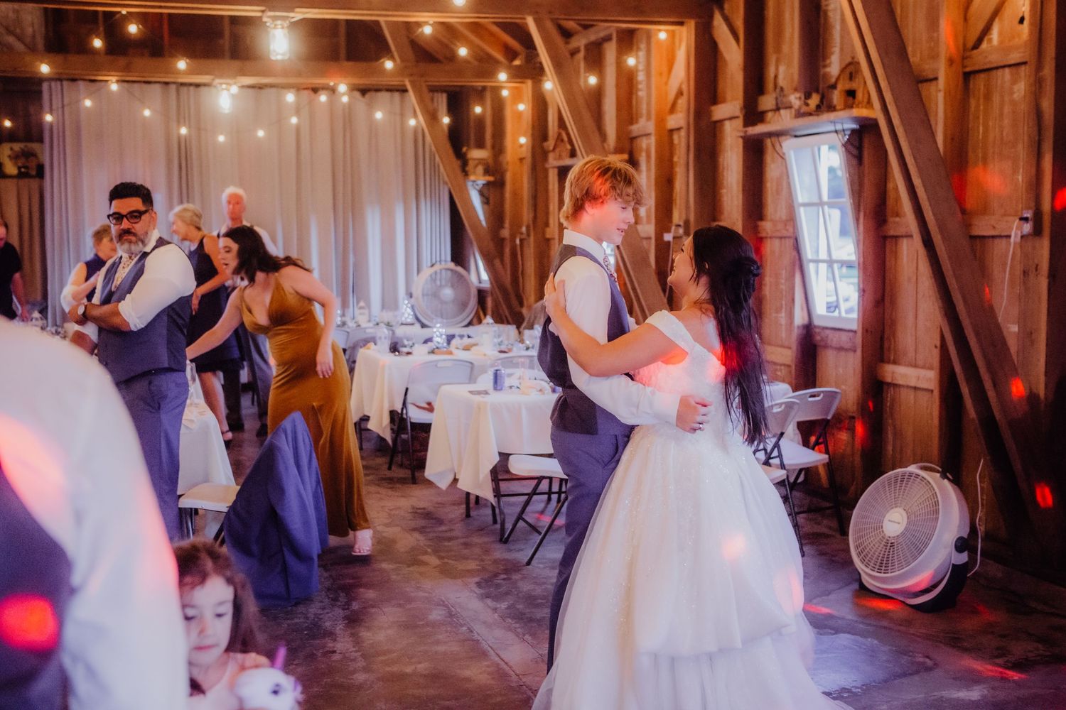 Wedding party dancing in a rustic barn decorated with string lights and exposed wooden beams.