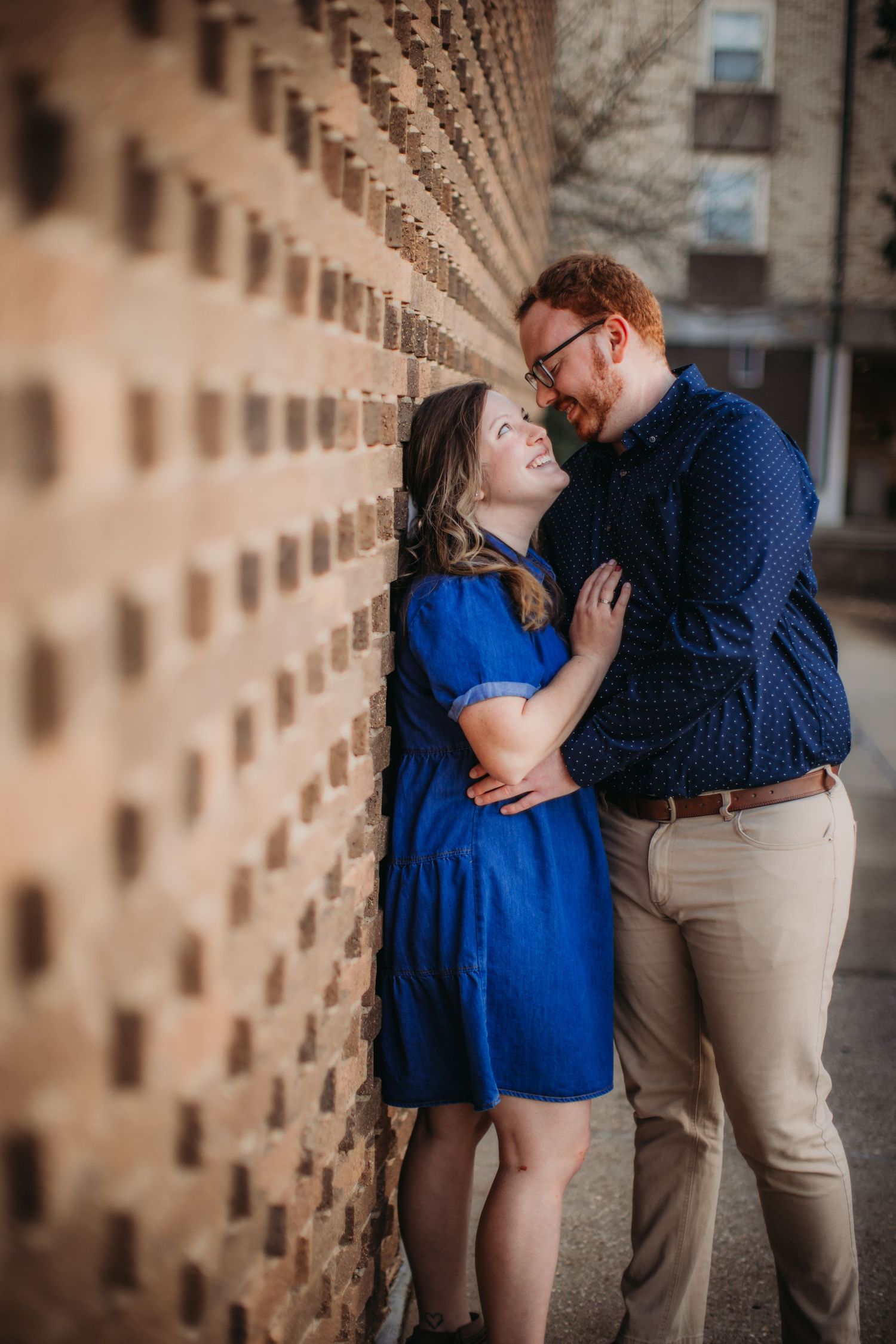 A romantic couple shares an intimate moment against a textured brick wall with repeating geometric patterns.