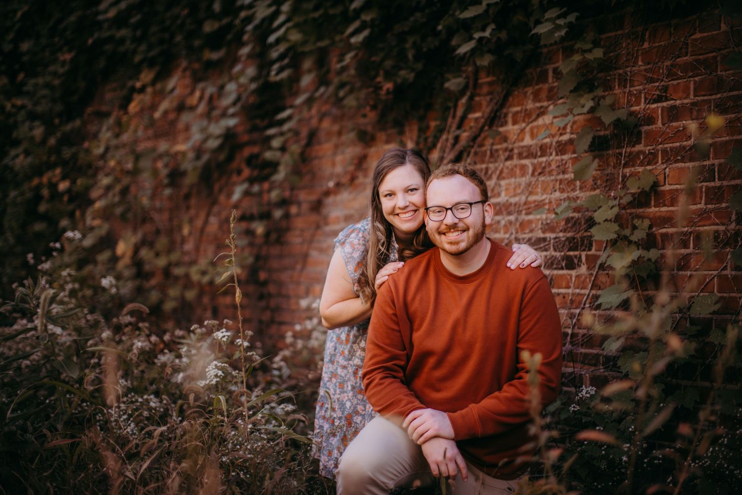 Sweet moment captured during an engagement session by a brick wall with natural foliage.