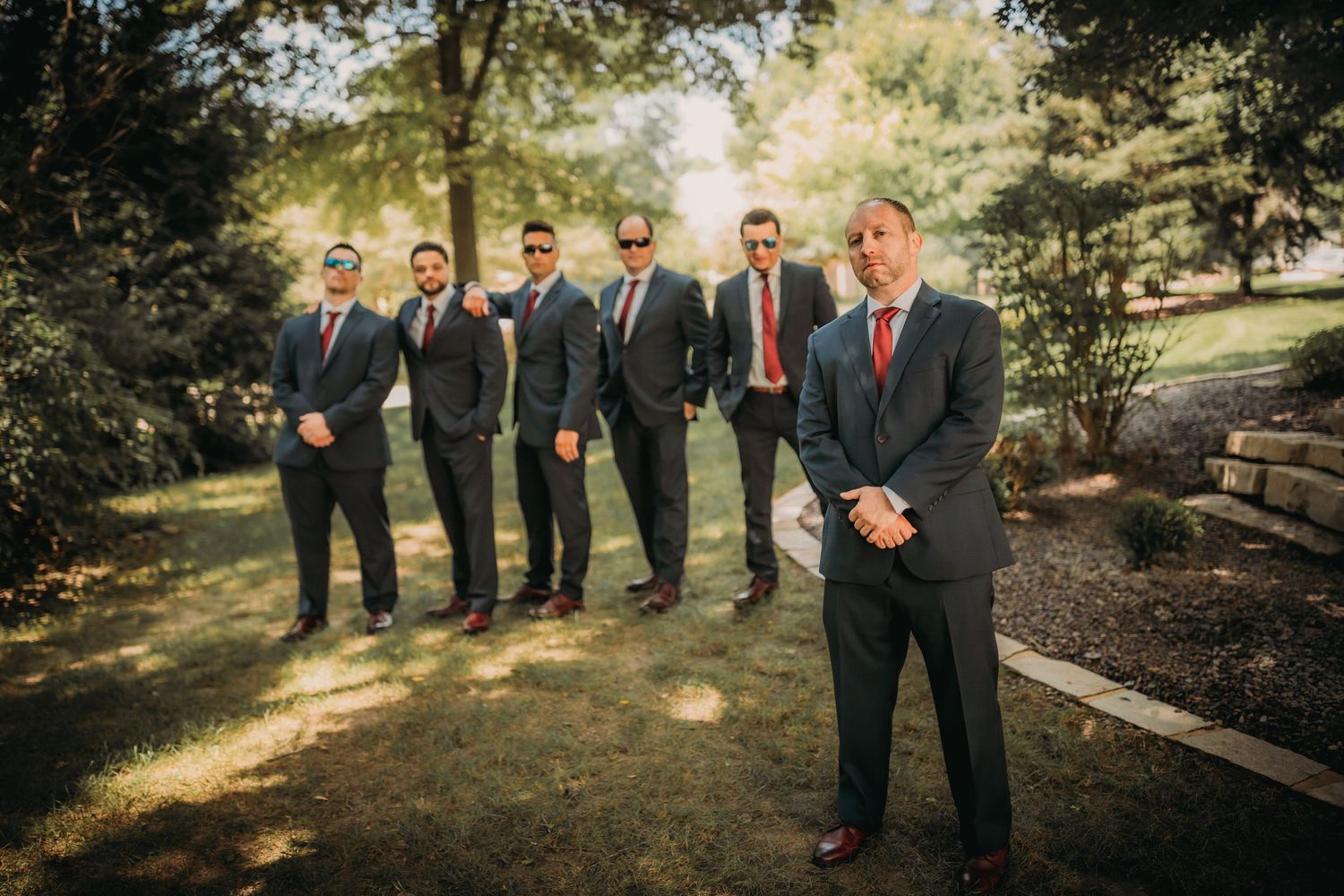 Groomsmen in dark suits and red ties stand in a garden setting during golden hour.