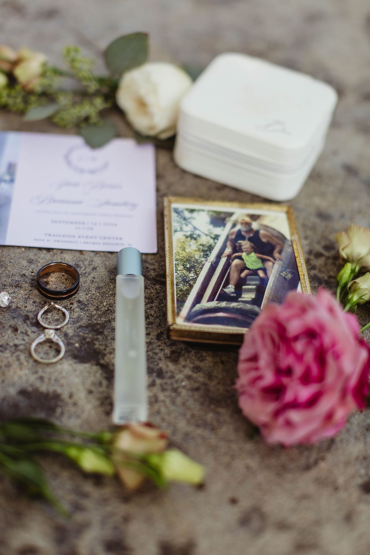 Wedding details including rings, photograph, invitation and pink rose on textured surface.