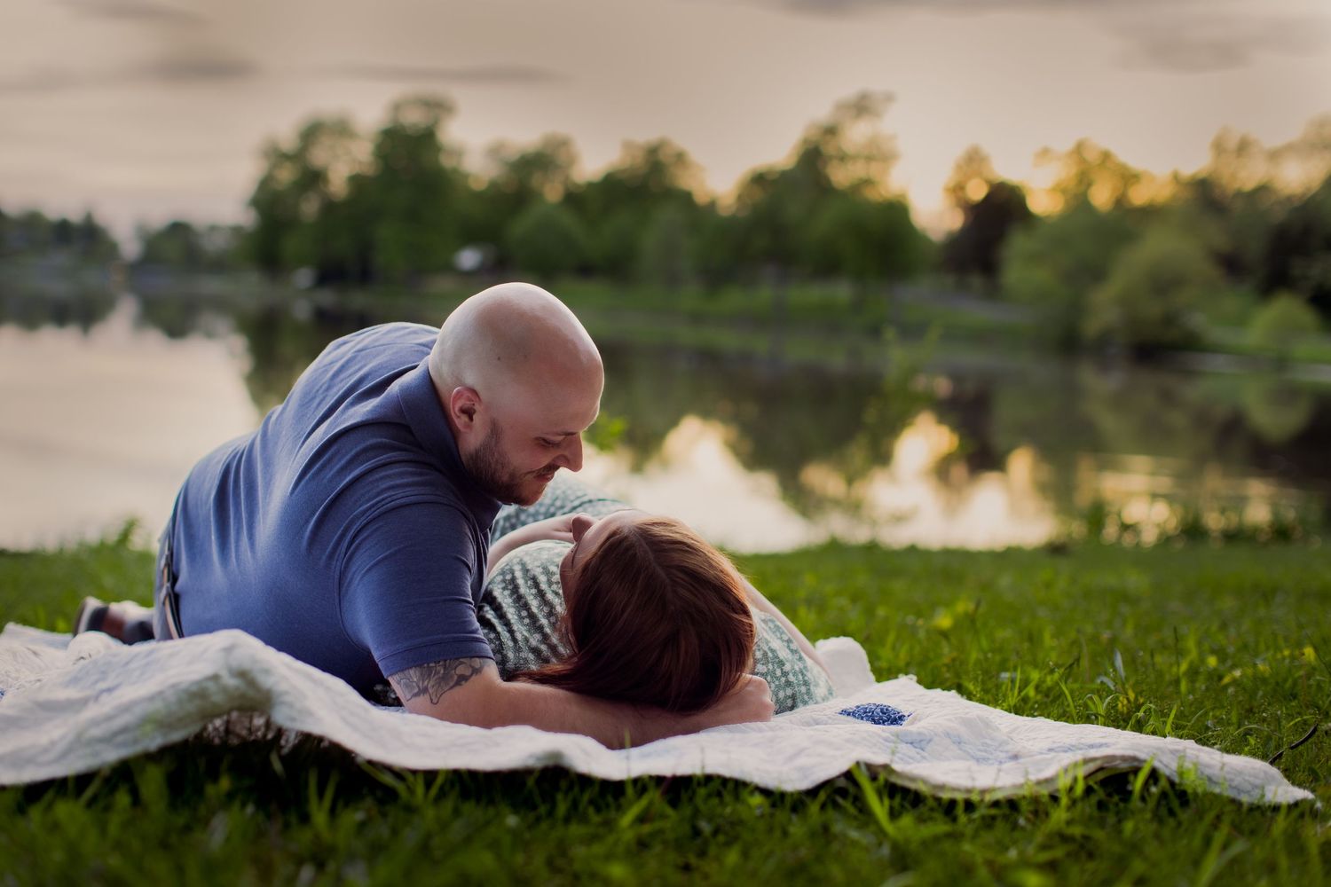 A couple laying on a white blanket by a lake during golden hour.