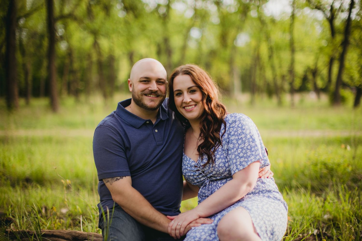 A couple poses together on a sunny day in a lush green field with trees in the background.