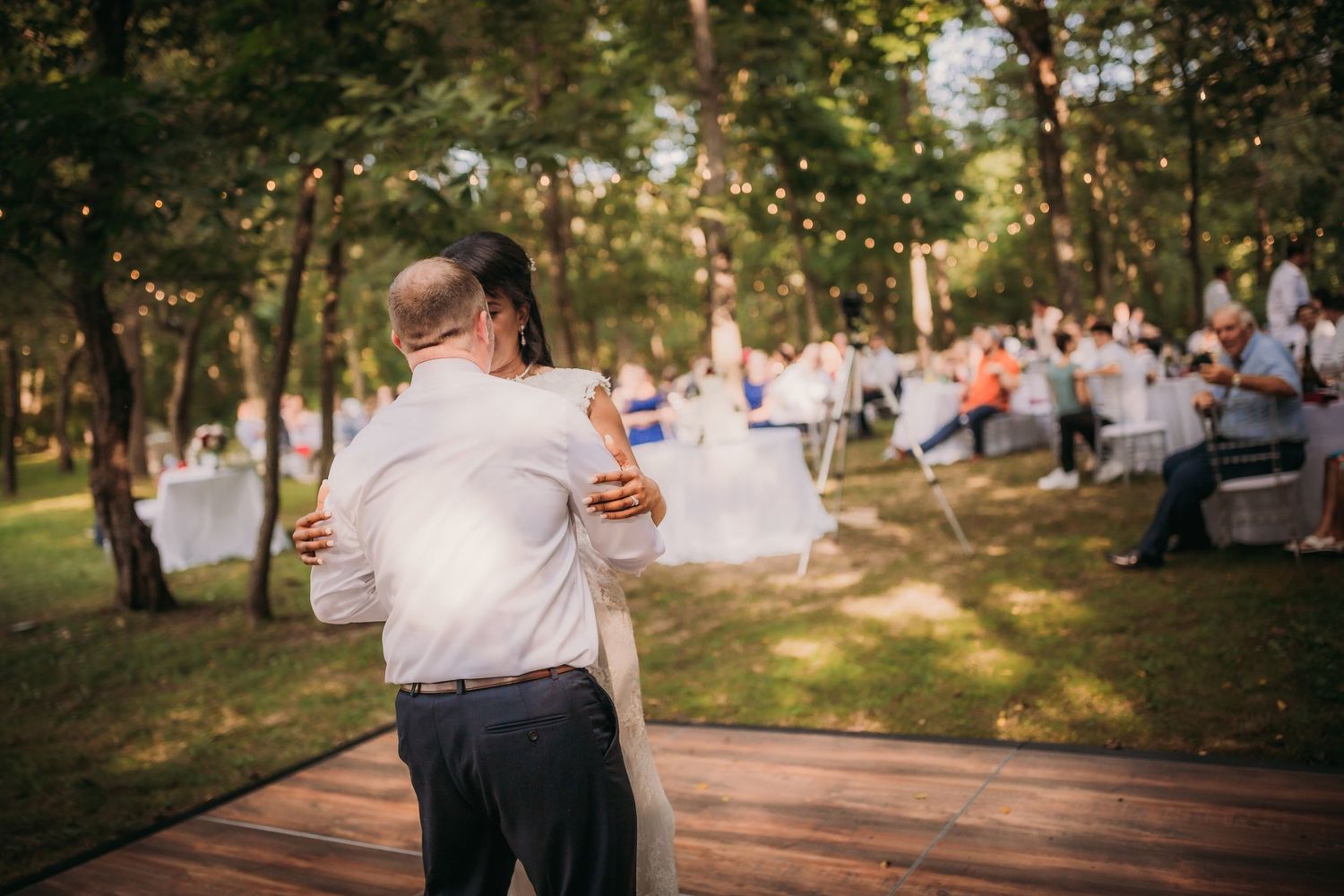 A couple dances at their outdoor wedding reception with wedding cake and guests visible in the background.